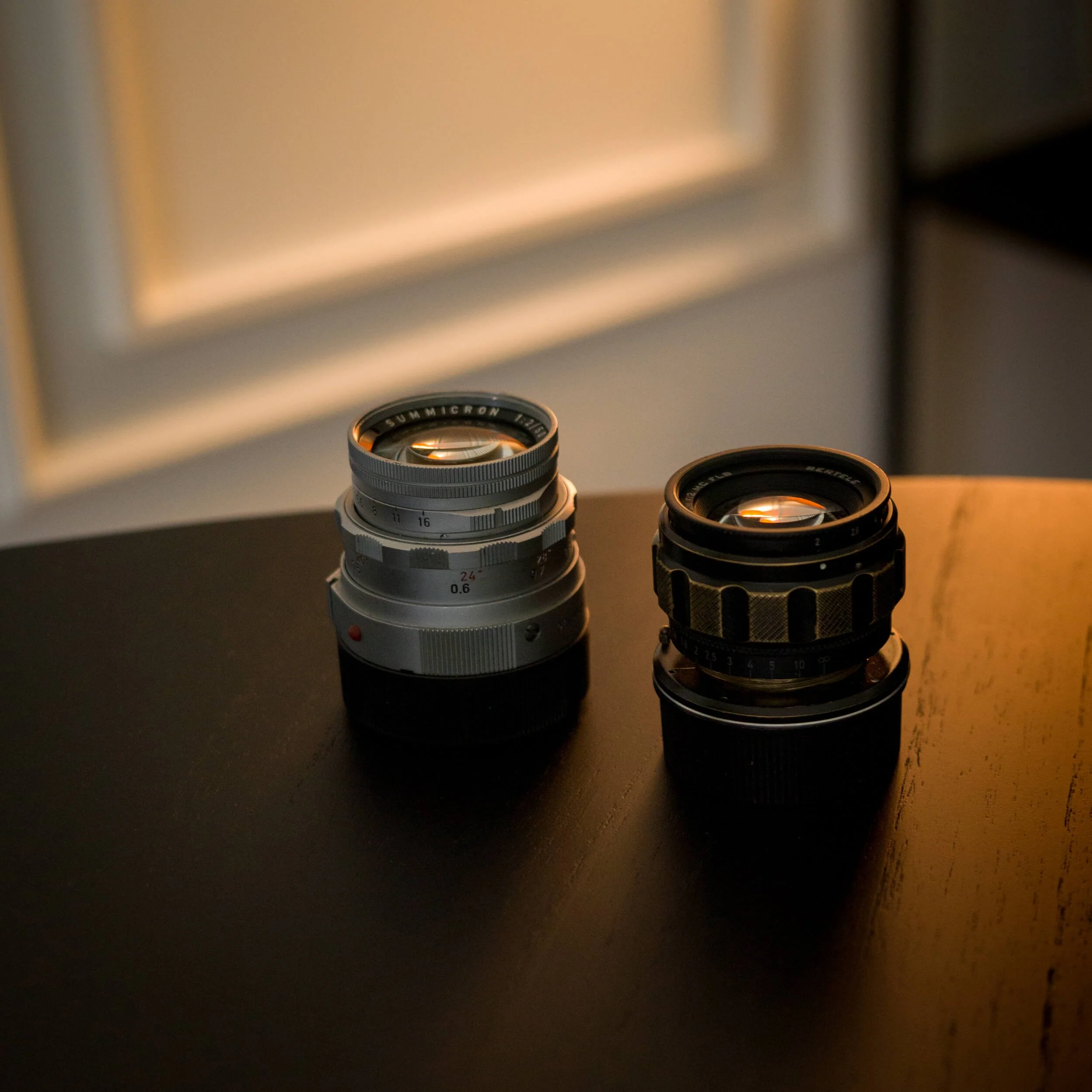 Product comparison between the Omnar Bertele 50mm f/2 and Leica Rigid Summicron 50mm lenses on a dark table