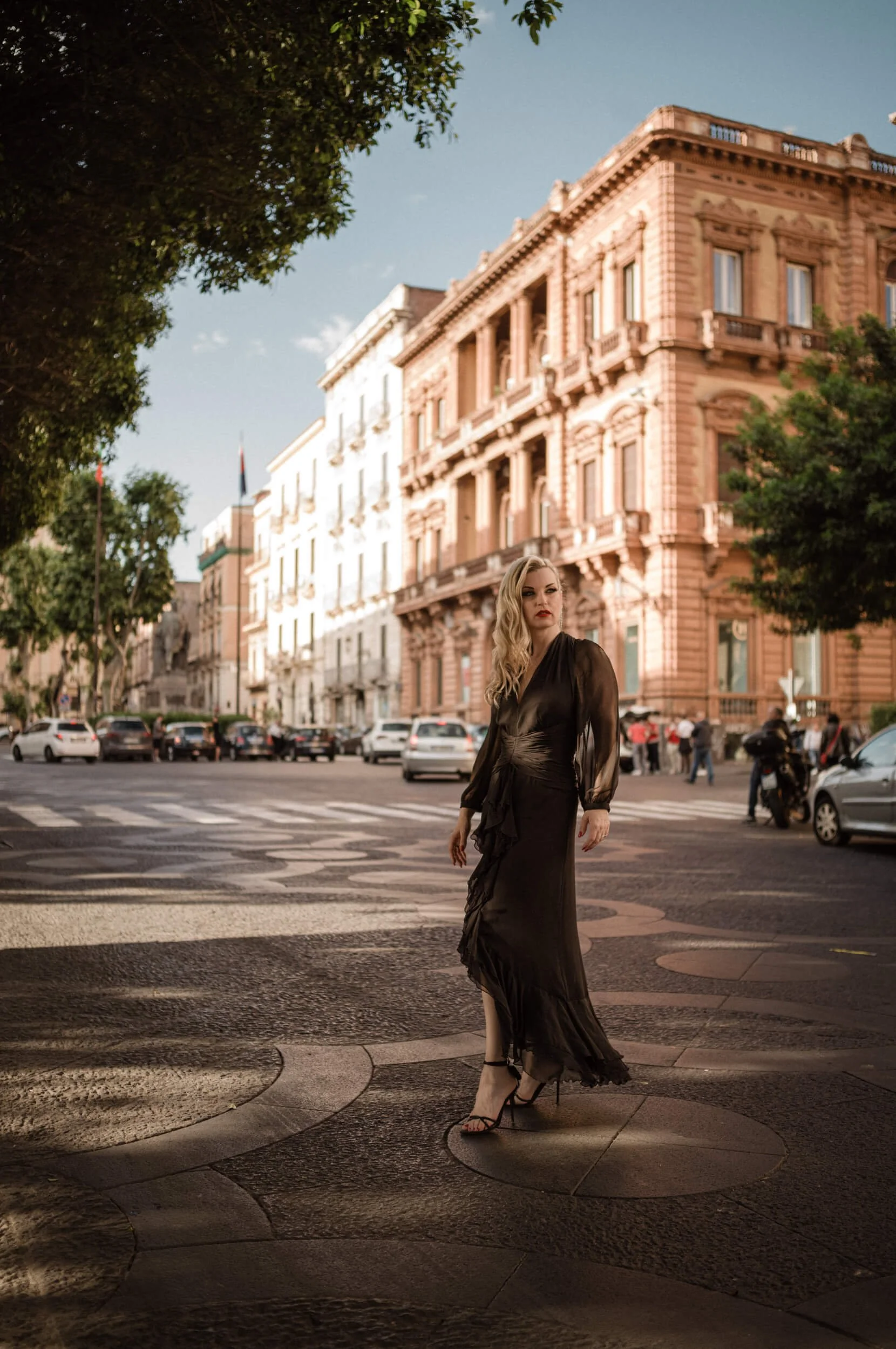 Roberta crossing a historic square in Catania, Sicily, wearing a flowing black dress during golden hour, photographed with the Thypoch Simera 35mm f/2.