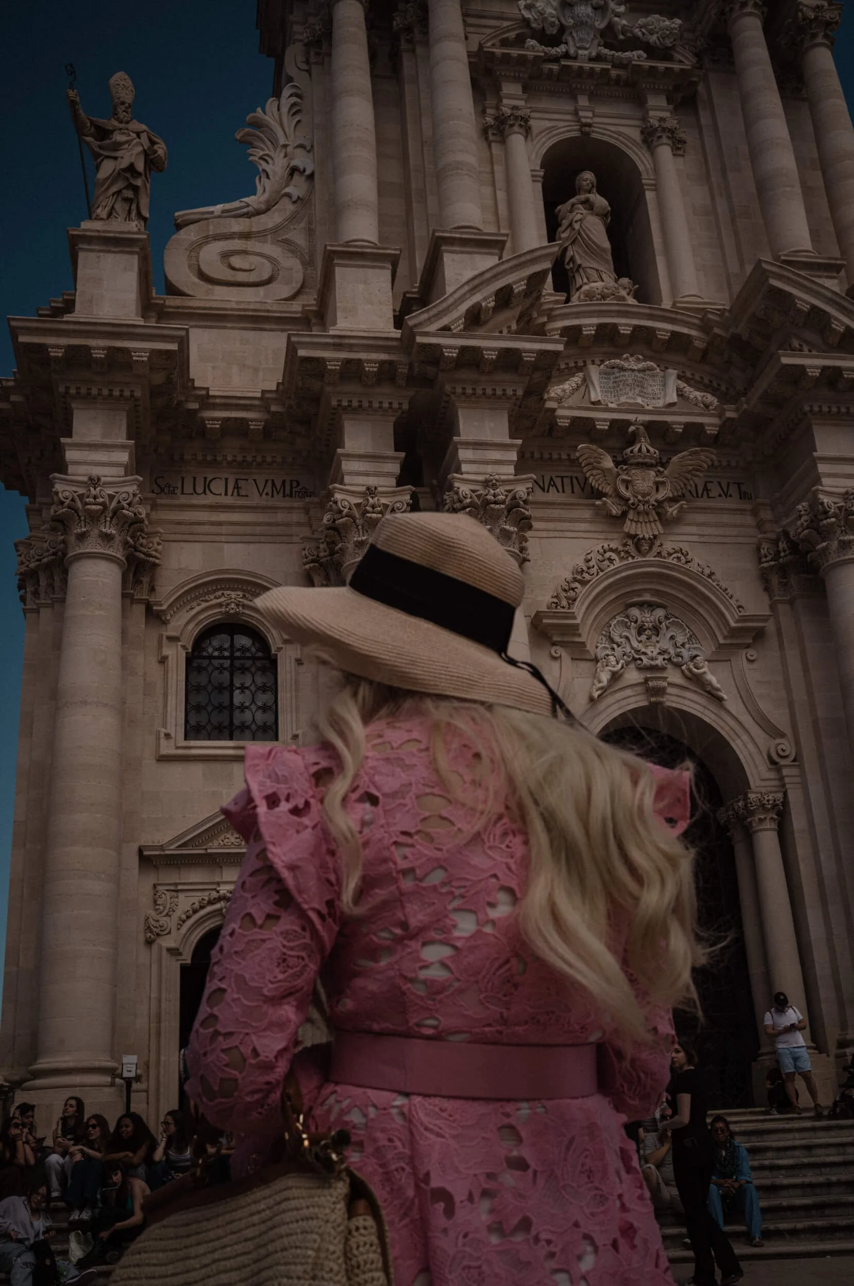 A tourist in a pink lace dress and straw hat standing before the Baroque façade of Syracuse Cathedral in Siracusa, Sicily, photographed with the Thypoch Simera 35mm f/2.