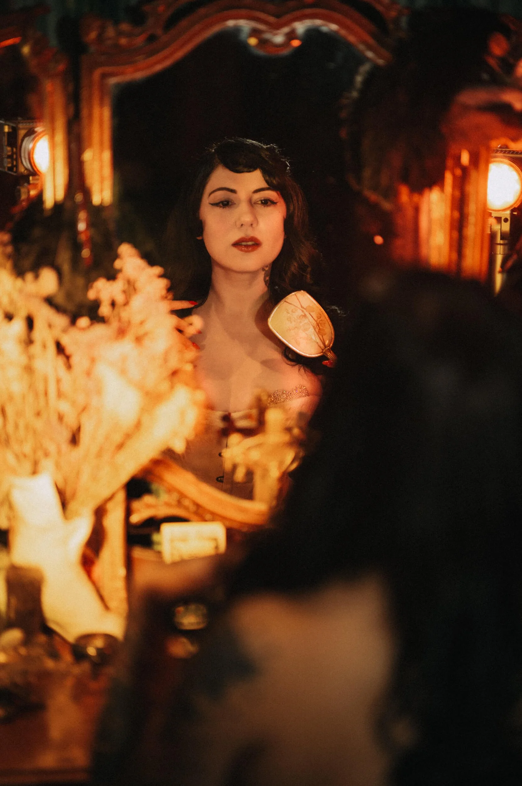 Bettie From Hell photographed at her vintage makeup table with the Thypoch Simera 75mm f/1.4 on the Leica M11-D, using warm red lighting and foreground reflections.