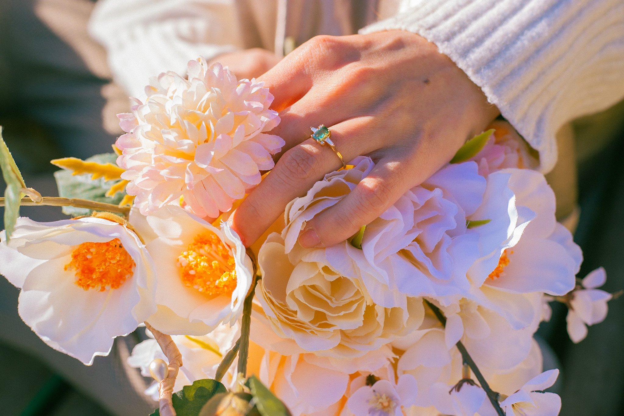 A person's hand resting on a bouquet of large, soft white and pale peach flowers, wearing a gold ring with a blue gemstone.