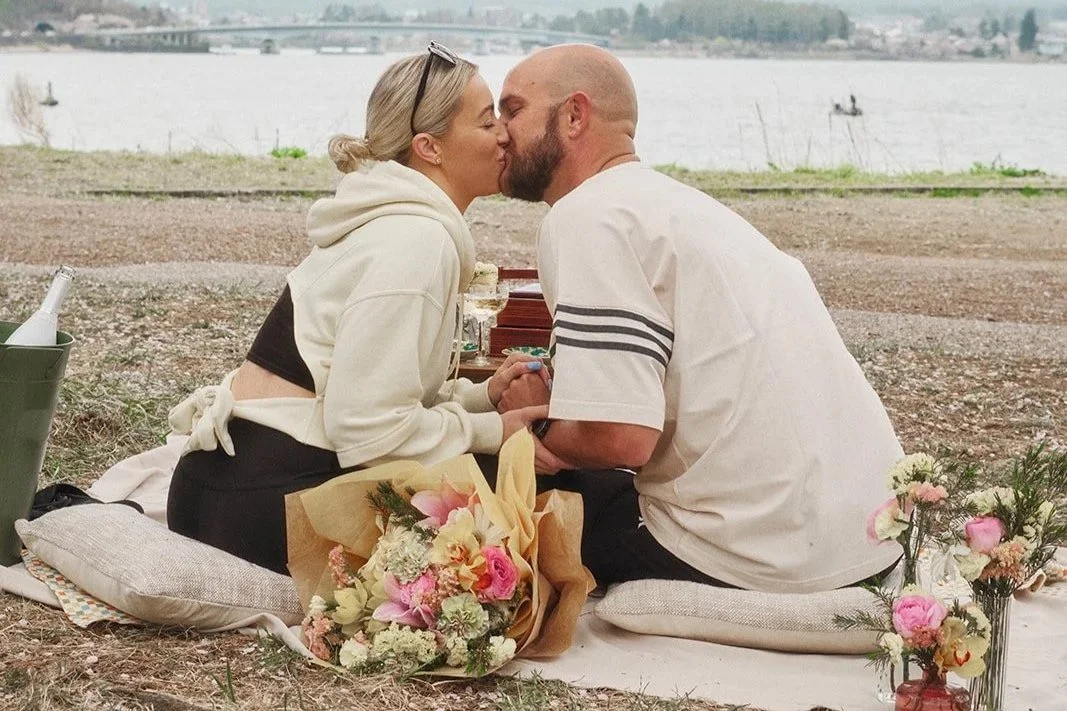 A couple kissing on a blanket during a picnic by the water, with flowers and bottles around them.