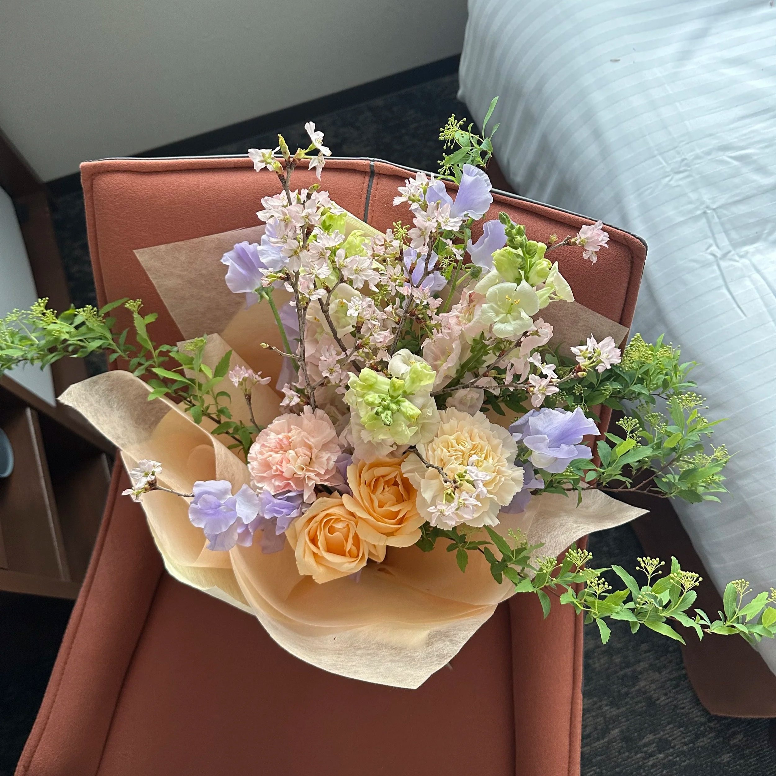 A colorful bouquet of flowers with peach roses, pink carnations, light purple delphiniums, and green foliage, wrapped in light-colored paper, placed on a dusty pink upholstered chair.