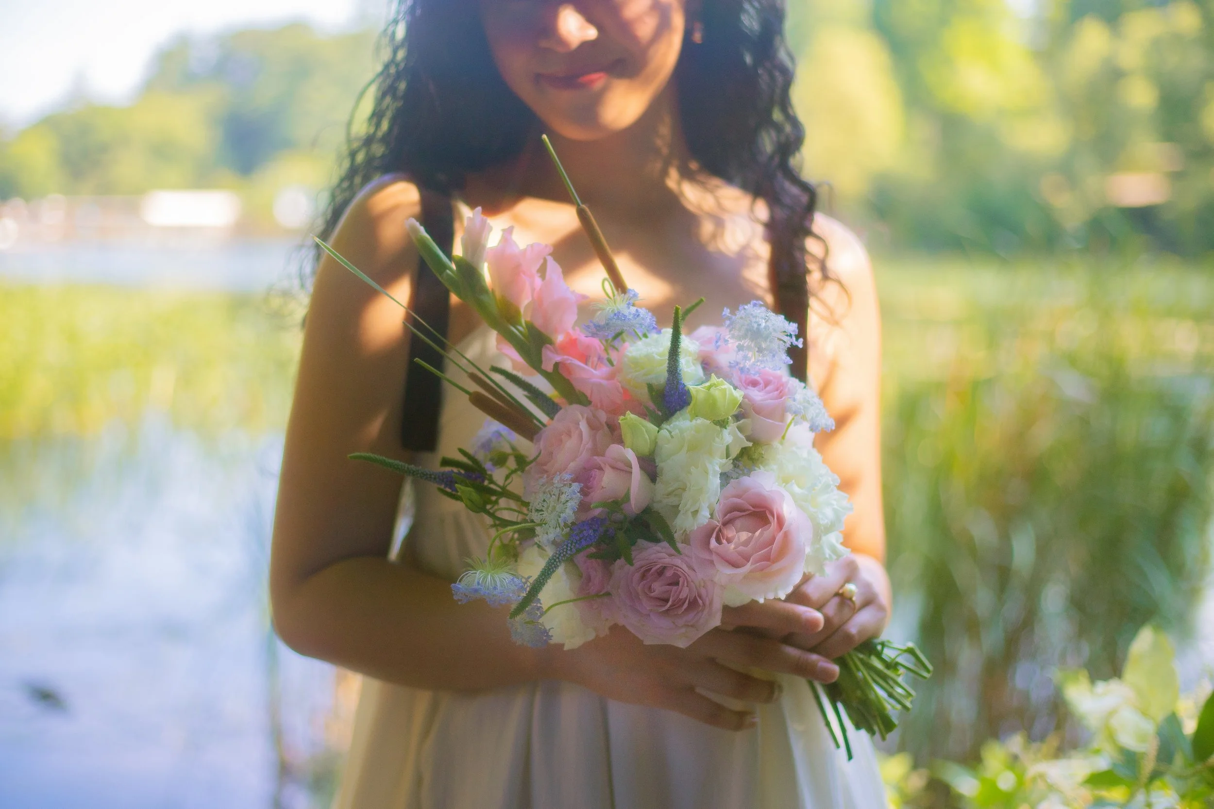 A woman holding a bouquet of pink, white, and purple flowers outdoors near a body of water, with trees and greenery in the background.