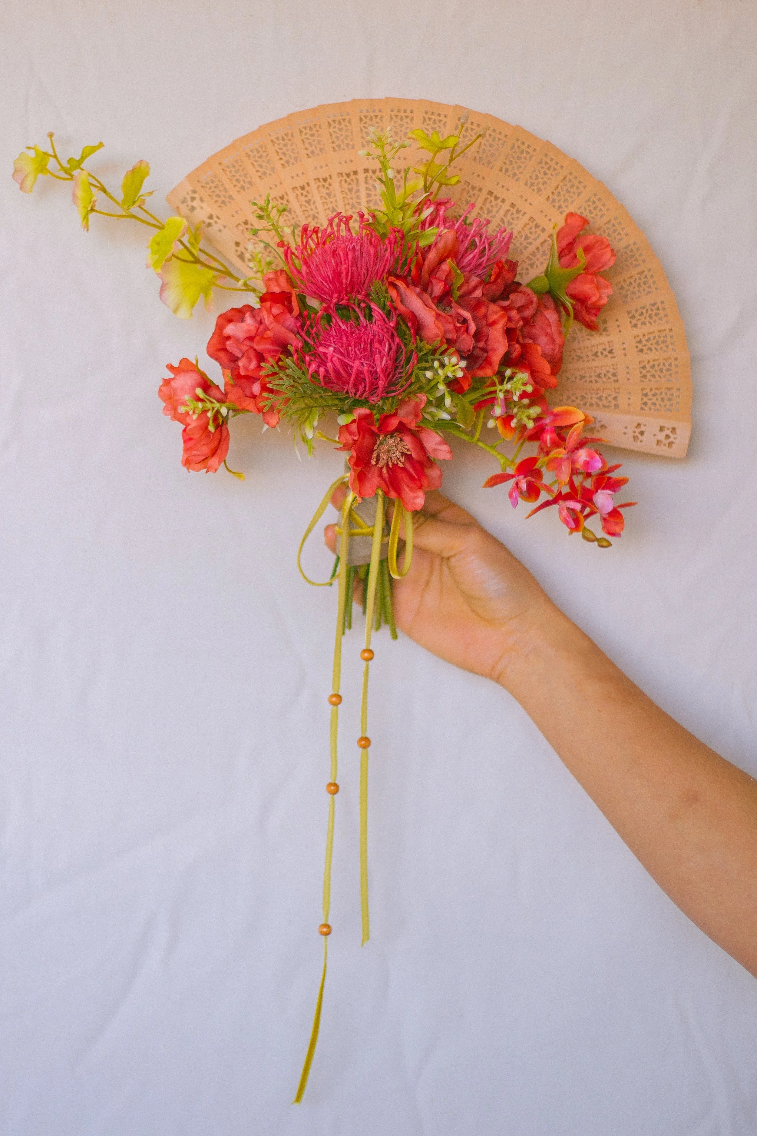A person's hand holding a bouquet of pink and orange flowers with a beige paper hand fan in the background.