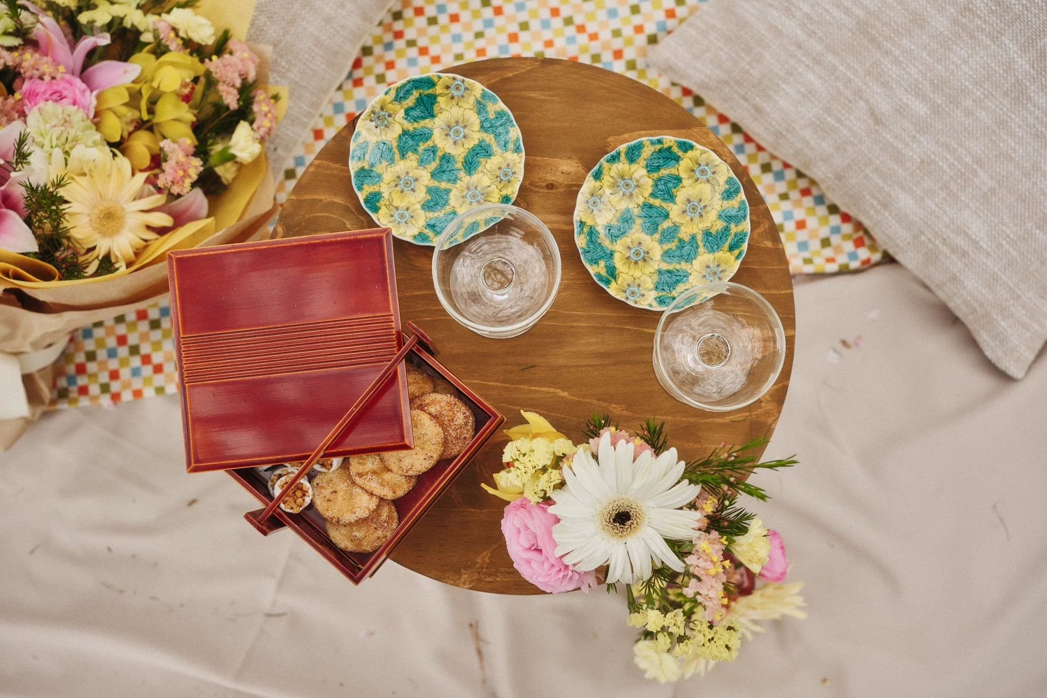 A top-down view of a table with two empty wine glasses, two floral-patterned plates, a stack of red square plates with cookies and chopsticks, a bouquet of pink, white, and yellow flowers, and a colorful patterned cloth in the background.