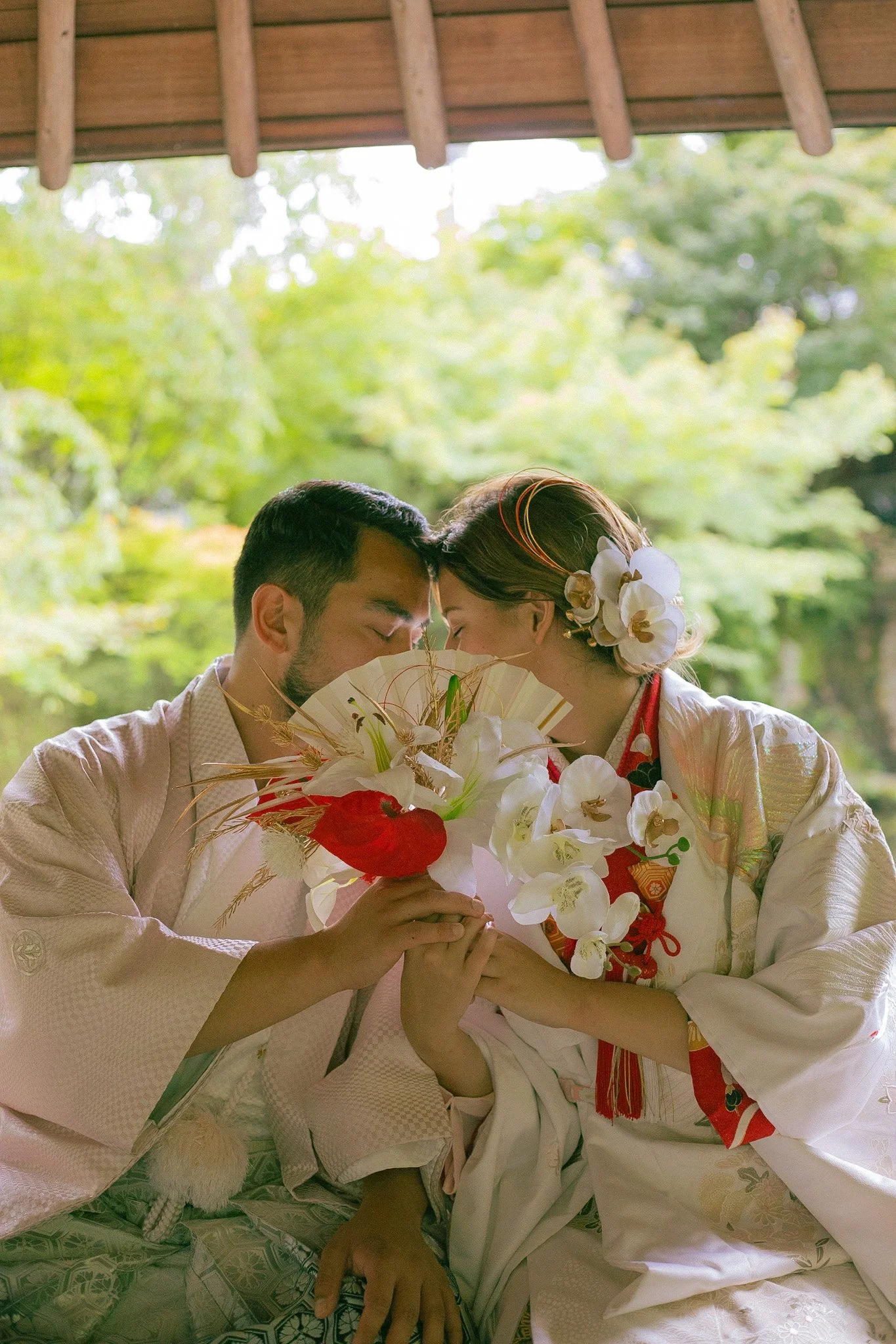A couple dressed in traditional Japanese wedding kimonos sharing a moment under a wooden gazebo with a lush green background, holding a bouquet of white orchids and red flowers.