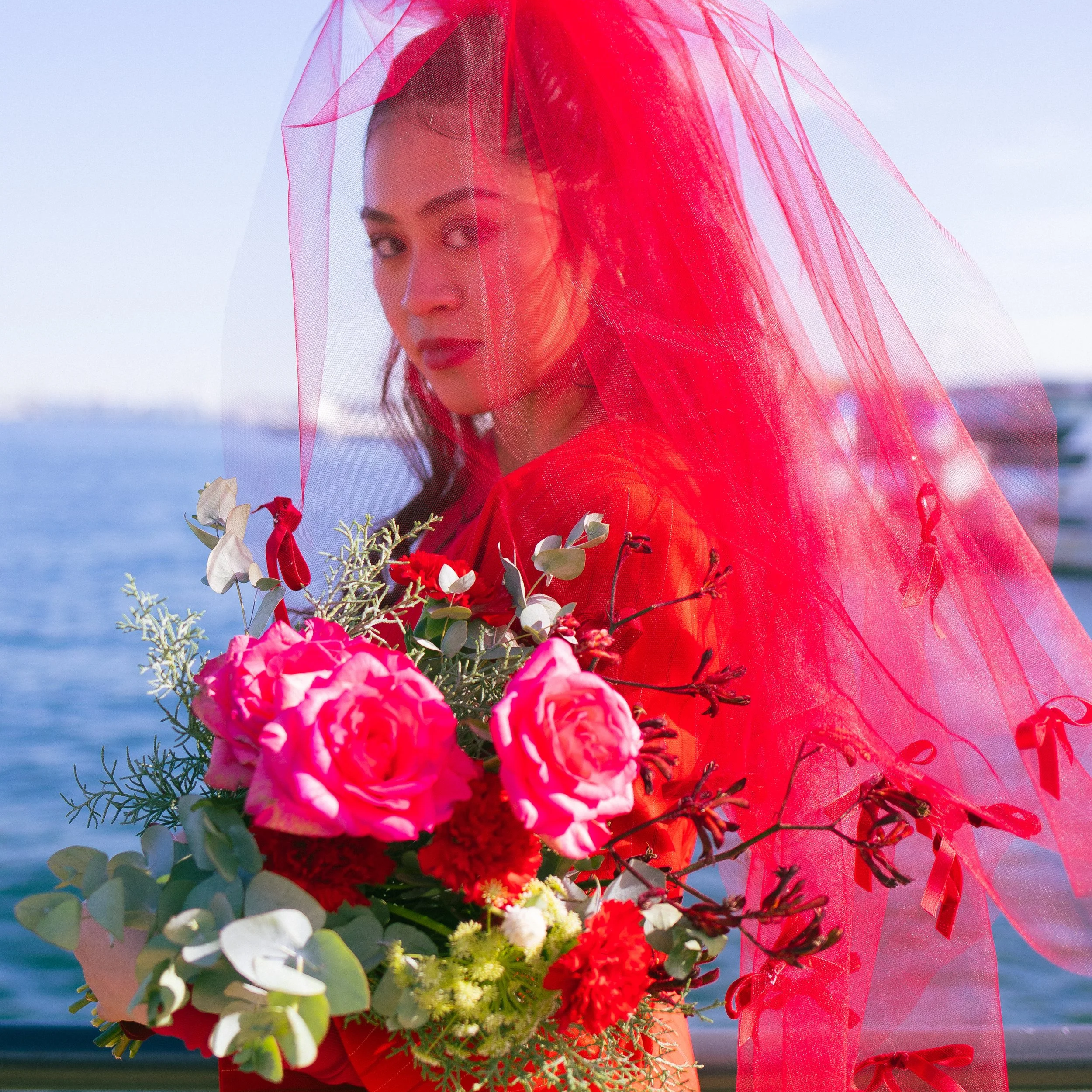 Woman wearing a red veil and dress, holding a bouquet of pink, red, and white flowers by the water.