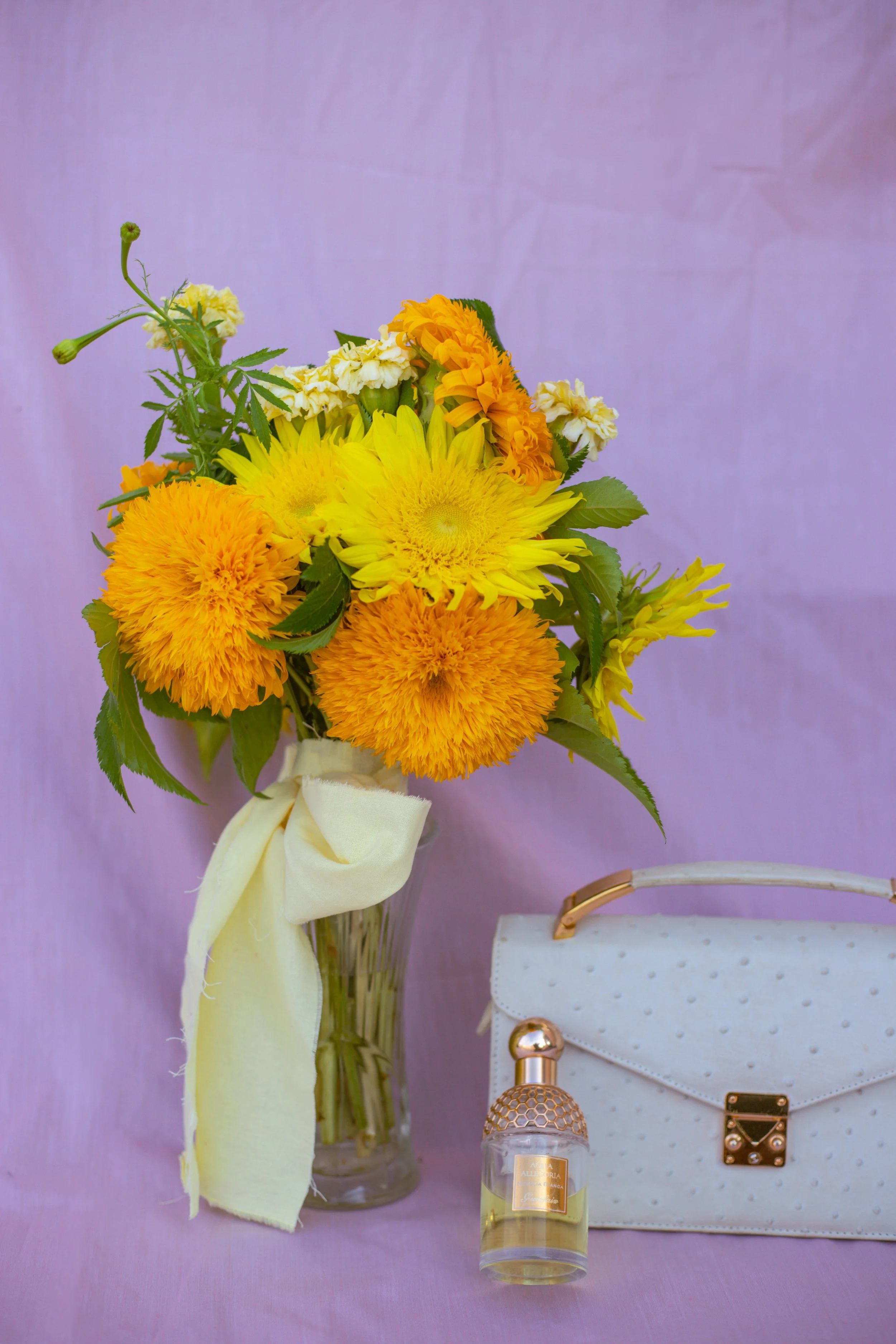 A bouquet of yellow and orange flowers in a glass vase, a white purse, and a small perfume bottle on a pink surface with a purple background.