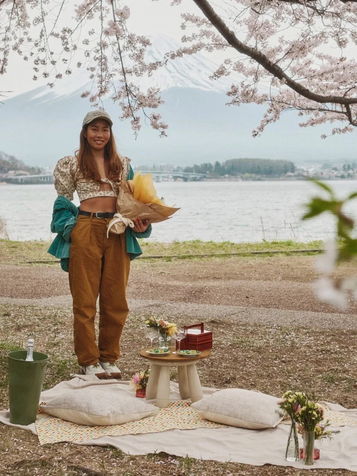 A woman holding a bouquet of flowers during a picnic with Mount Fuji in the background, cherry blossom trees, and a lake.