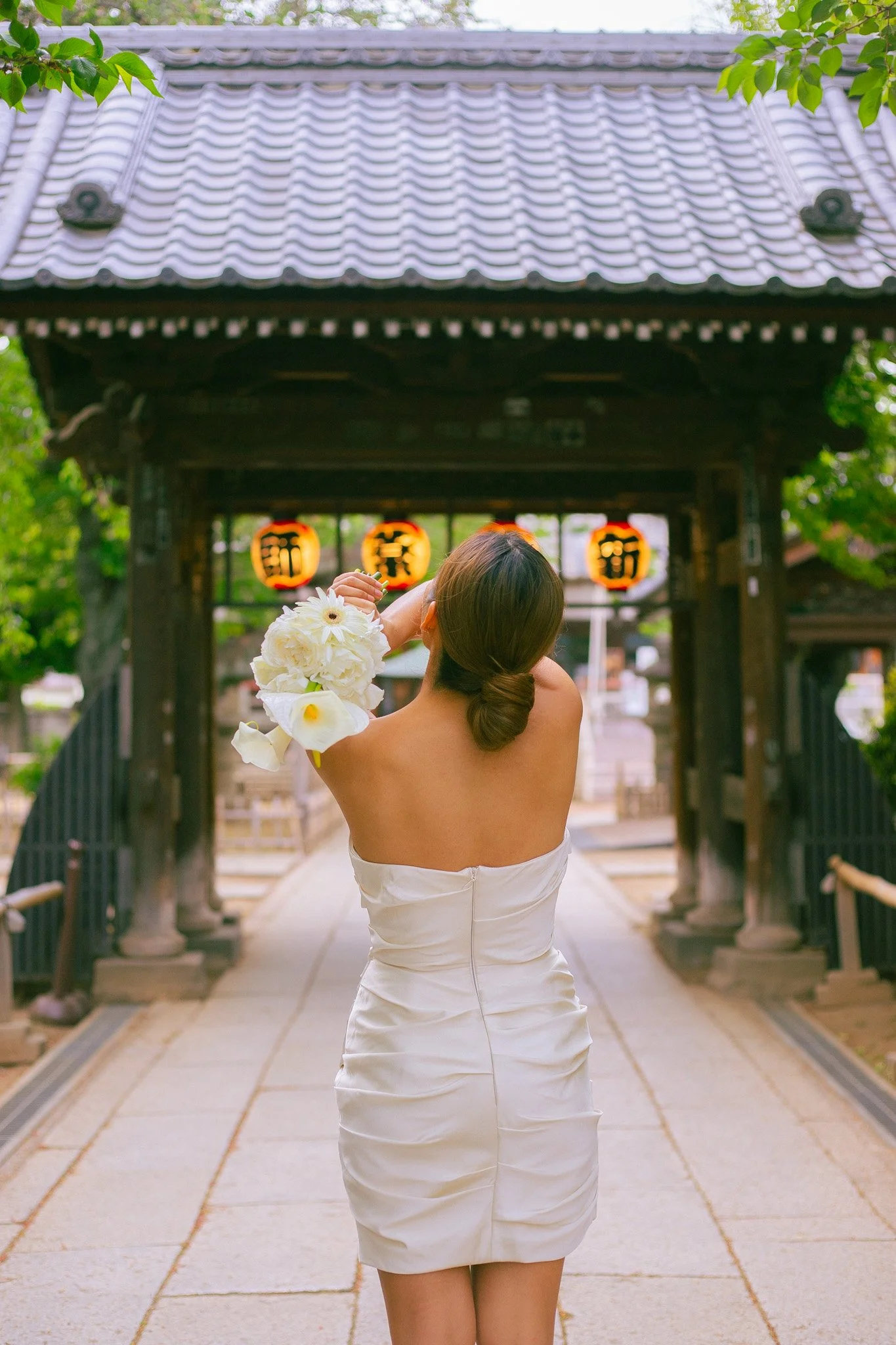 A woman in a white strapless dress holds a bouquet of white flowers, standing with her back to the camera, in front of a traditional Japanese gate with lanterns hanging inside.