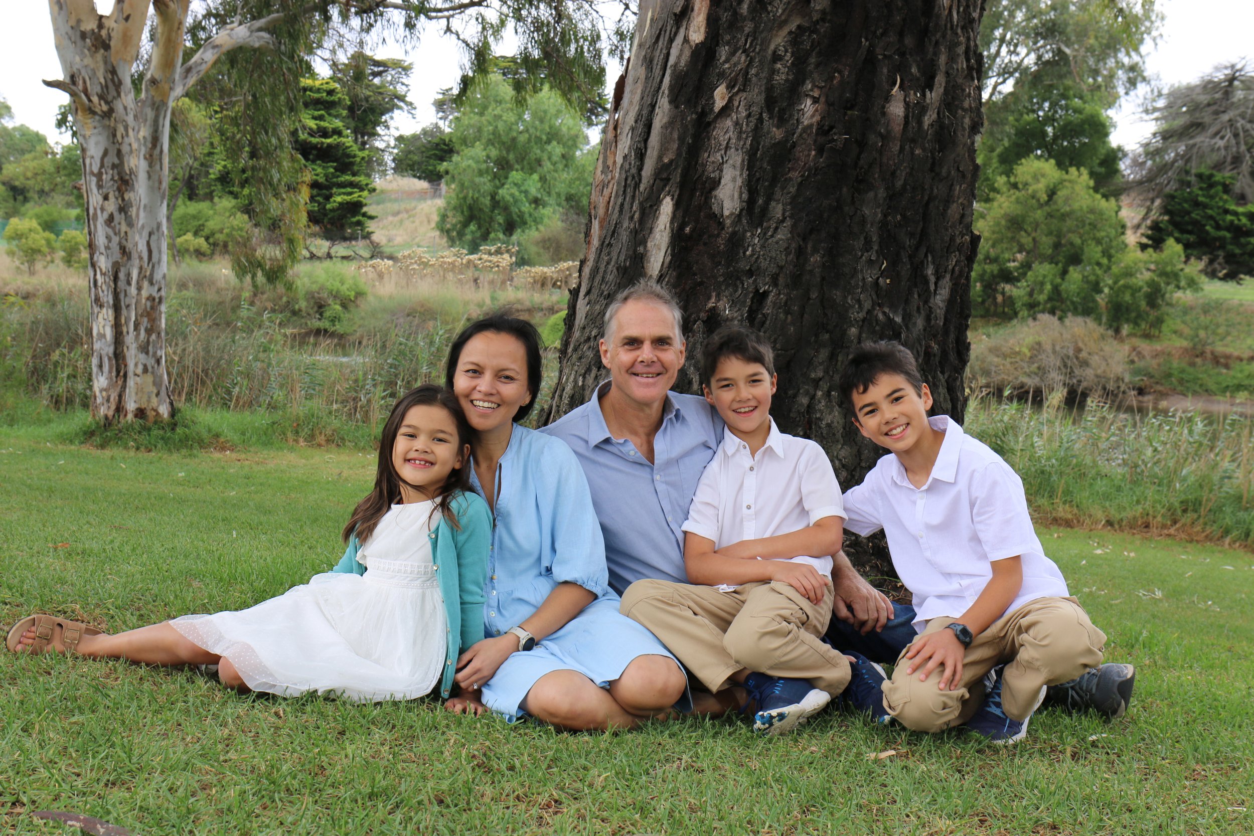 Family sitting on grass by a tree, smiling, outdoors