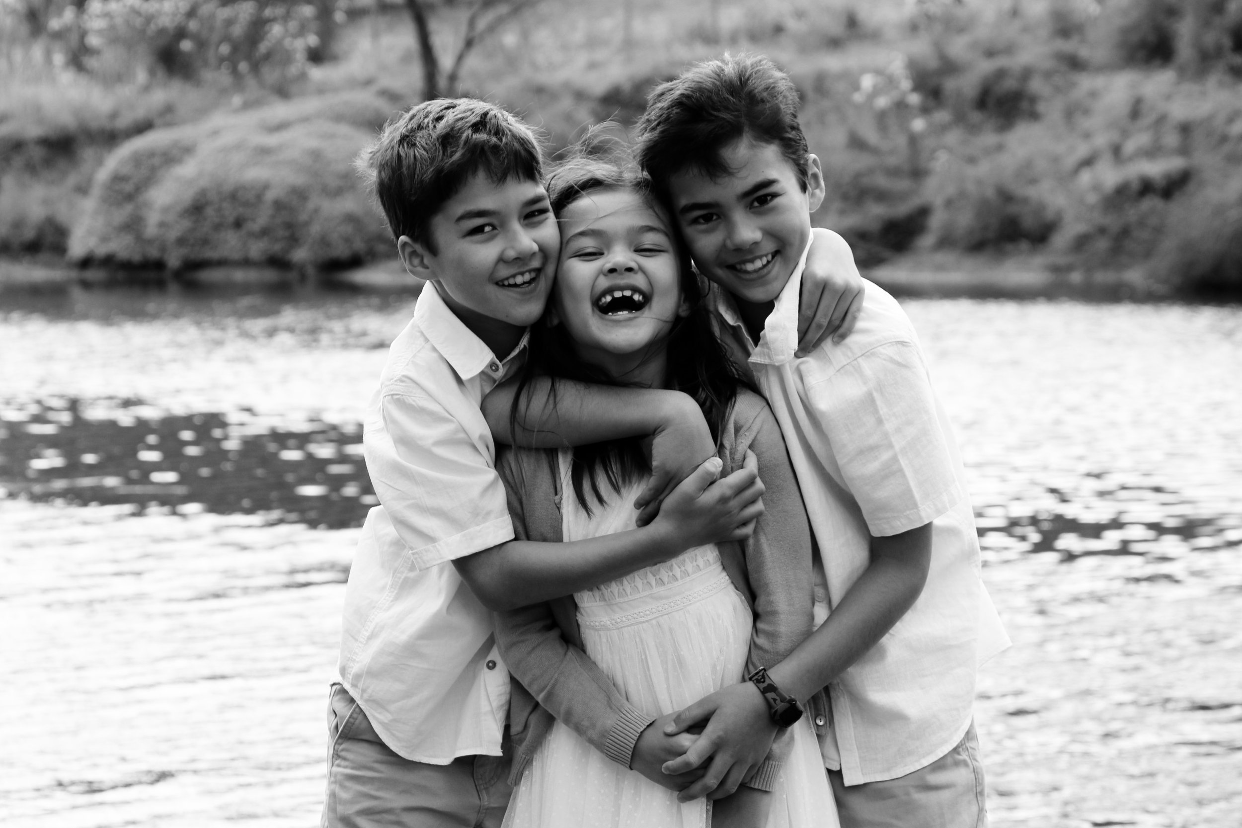 Three smiling children standing by a river, hugging and laughing, in a black and white photograph.