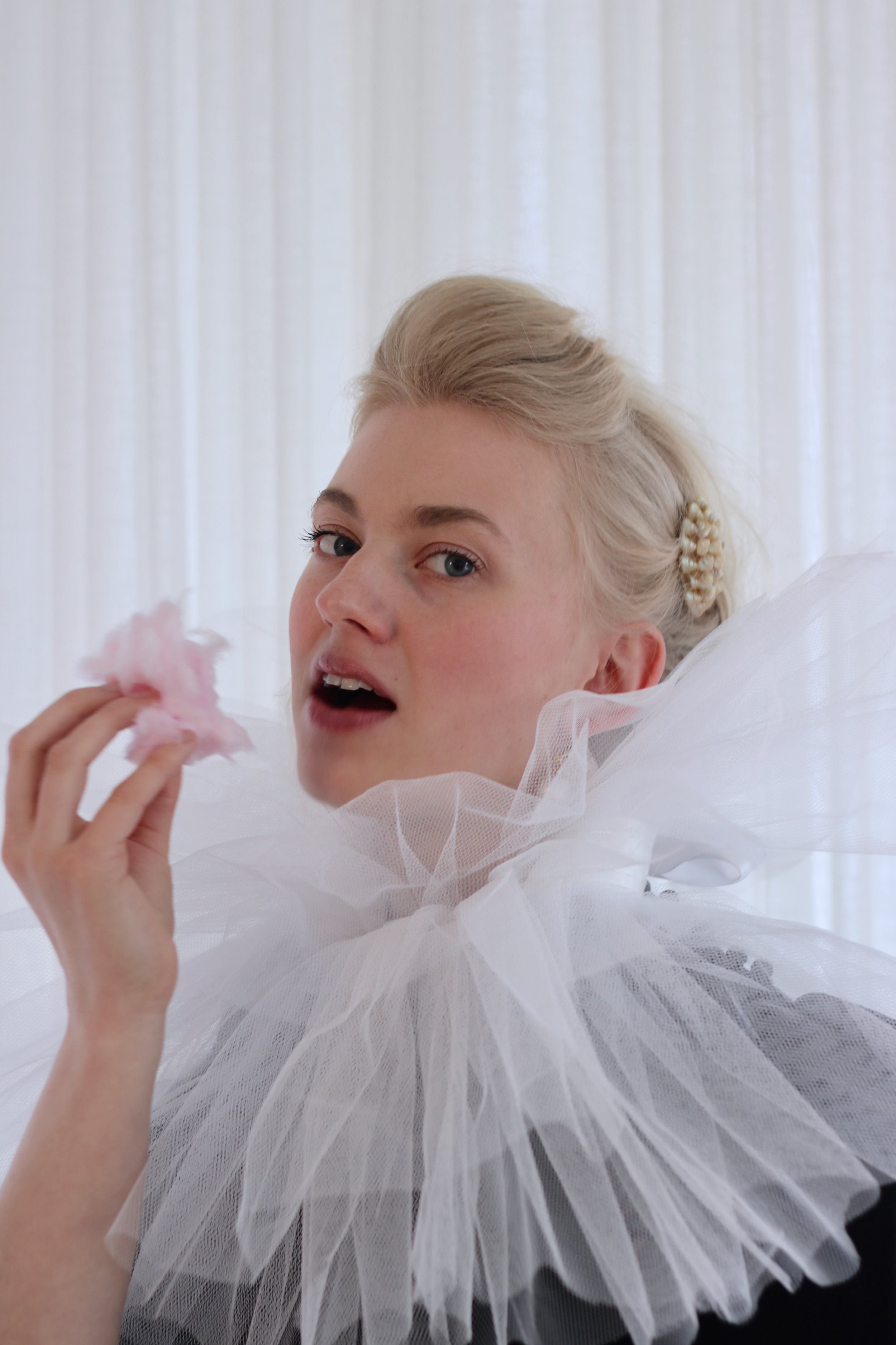 Person with a blonde hairstyle wearing a dramatic white tulle collar, holding a pink flower-like object near their mouth, set against a white background.