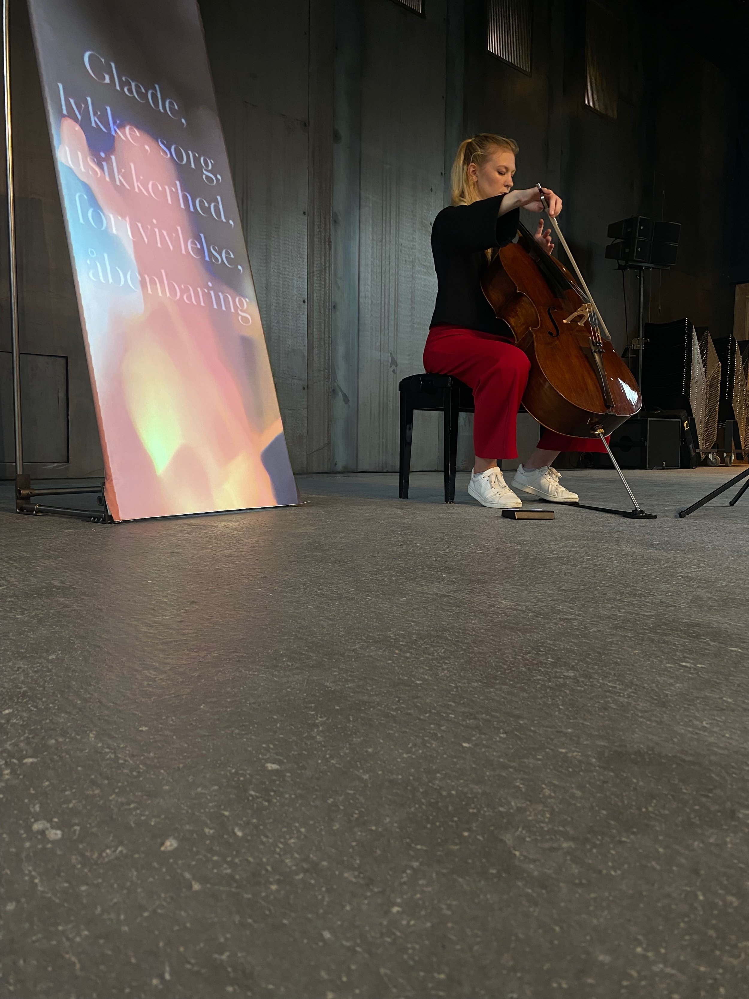 Woman playing cello on stage with a colorful sign displaying Danish words in the background.