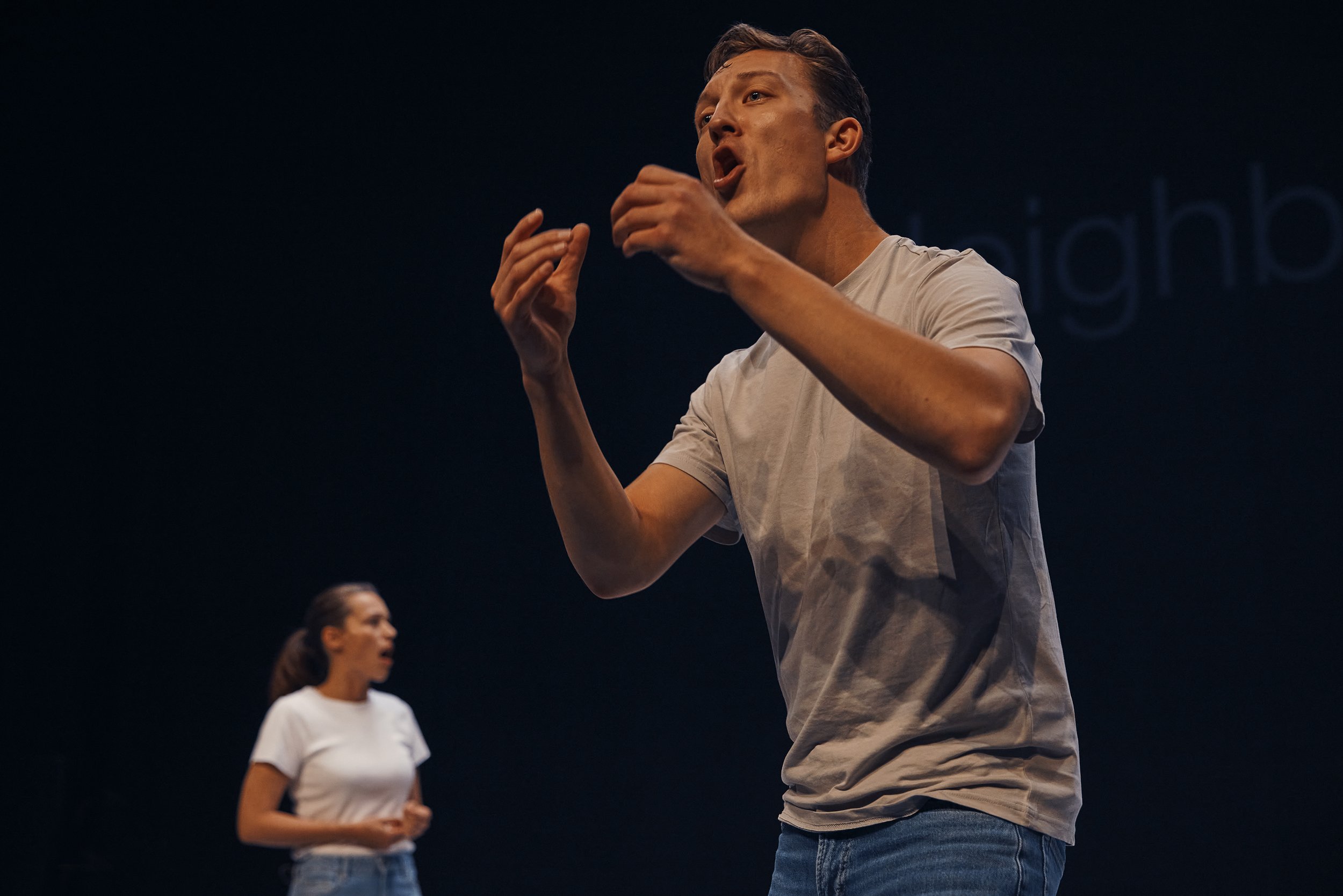 Two people performing on stage, wearing casual clothes; one person in the foreground gesturing passionately, the other in the background also engaged.