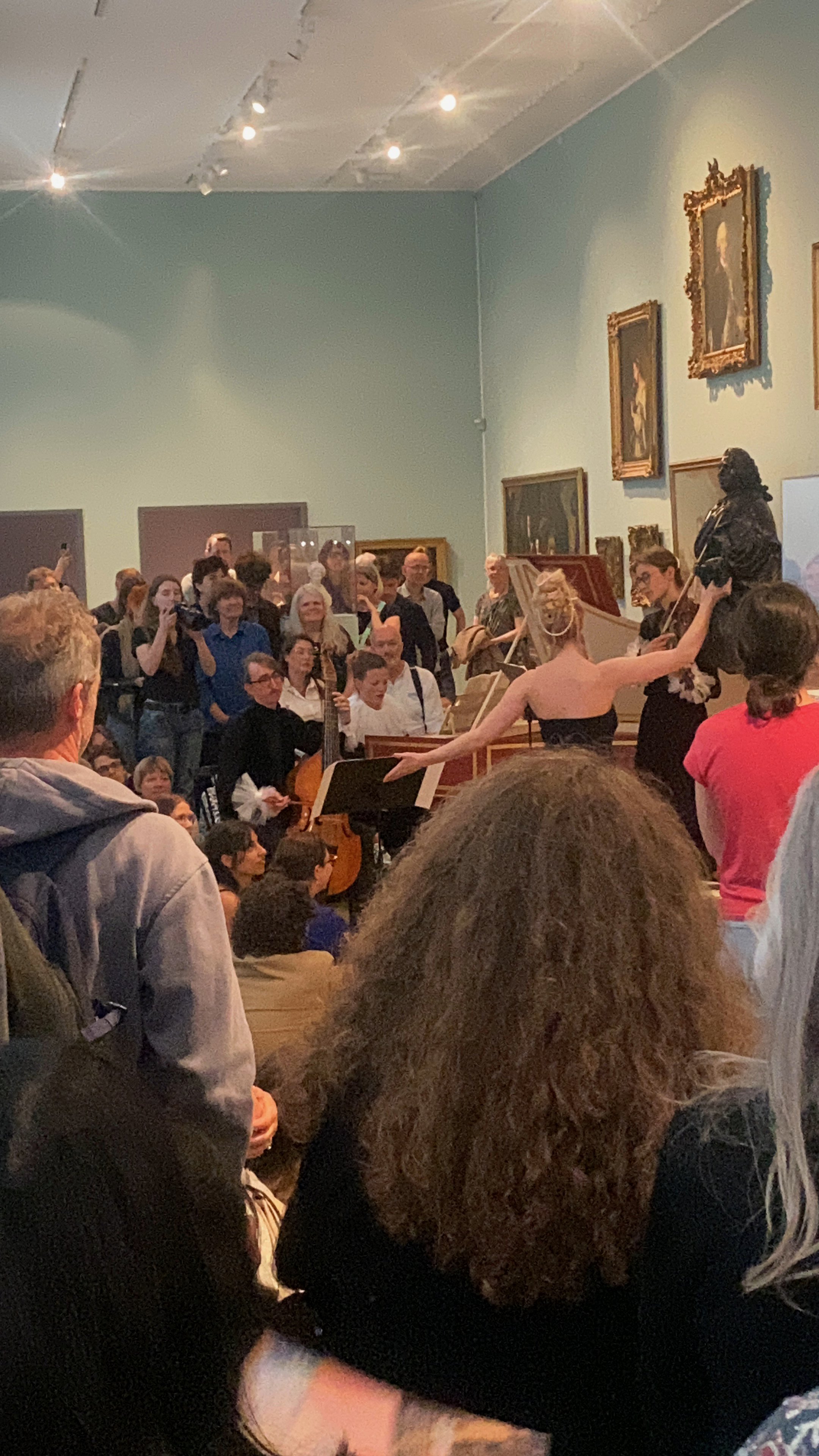 A group of people gathered indoors, some standing and some sitting, watching a performance with musicians and a person dancing. There are paintings and a sculpture on the wall.