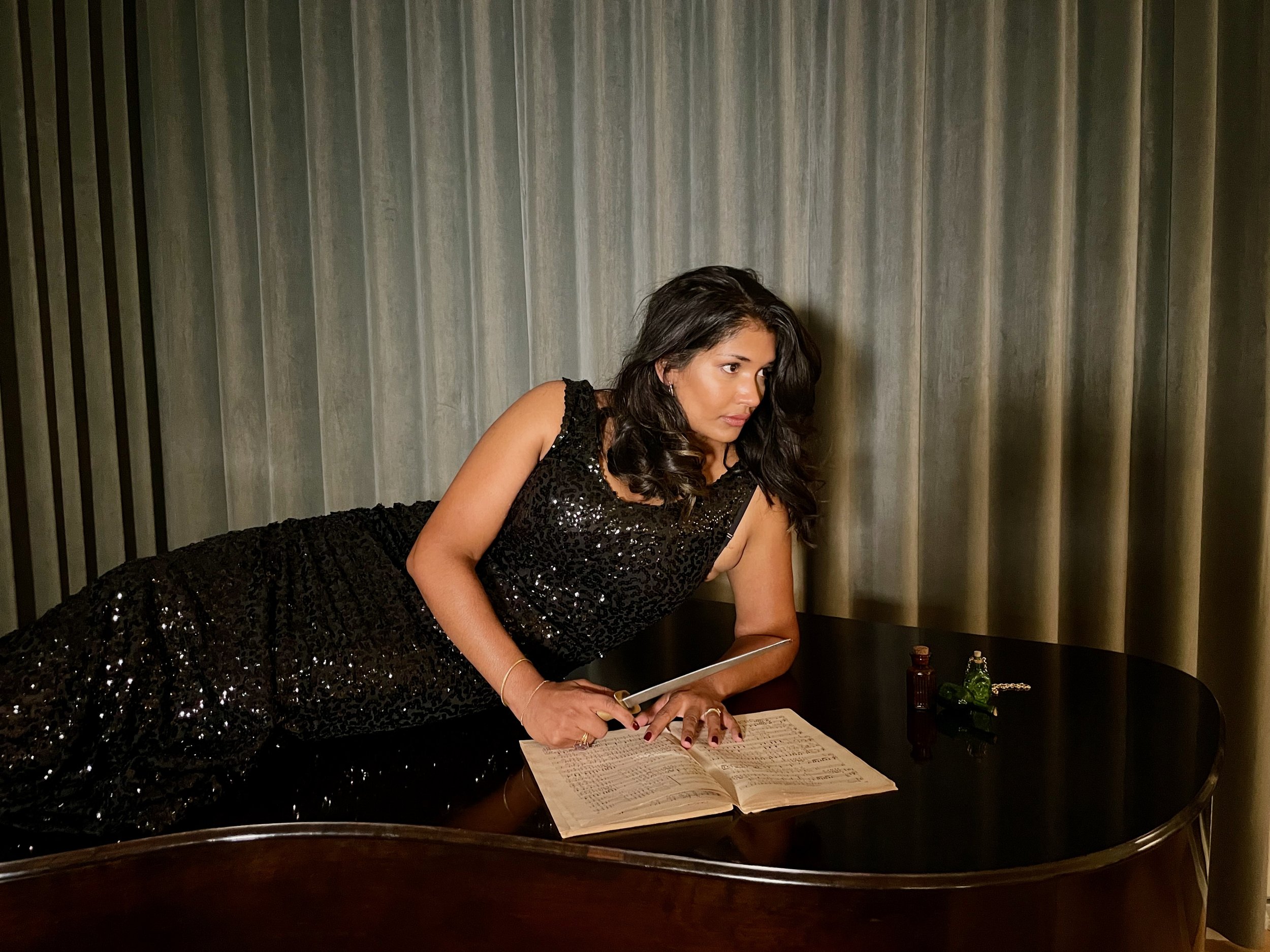 Woman in a black sequined dress lying on a grand piano with sheet music and small decorative items on the piano surface, against a curtain backdrop.