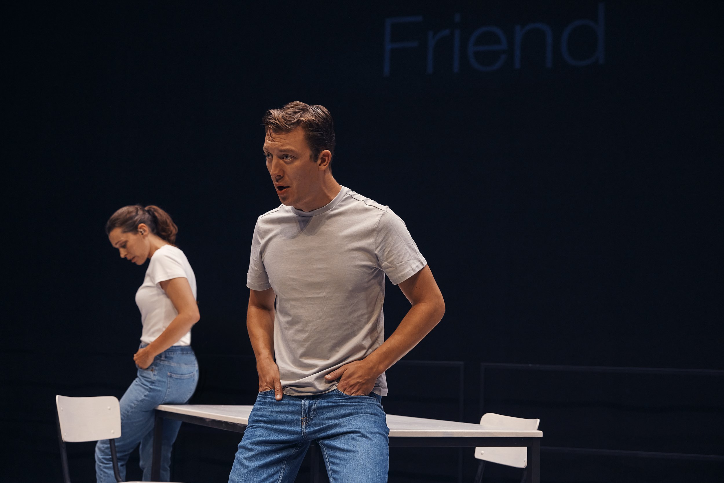 Two people, a man and a woman, wearing white t-shirts and jeans, are performing on stage with a table and chairs. The word "Friend" is visible in the background.