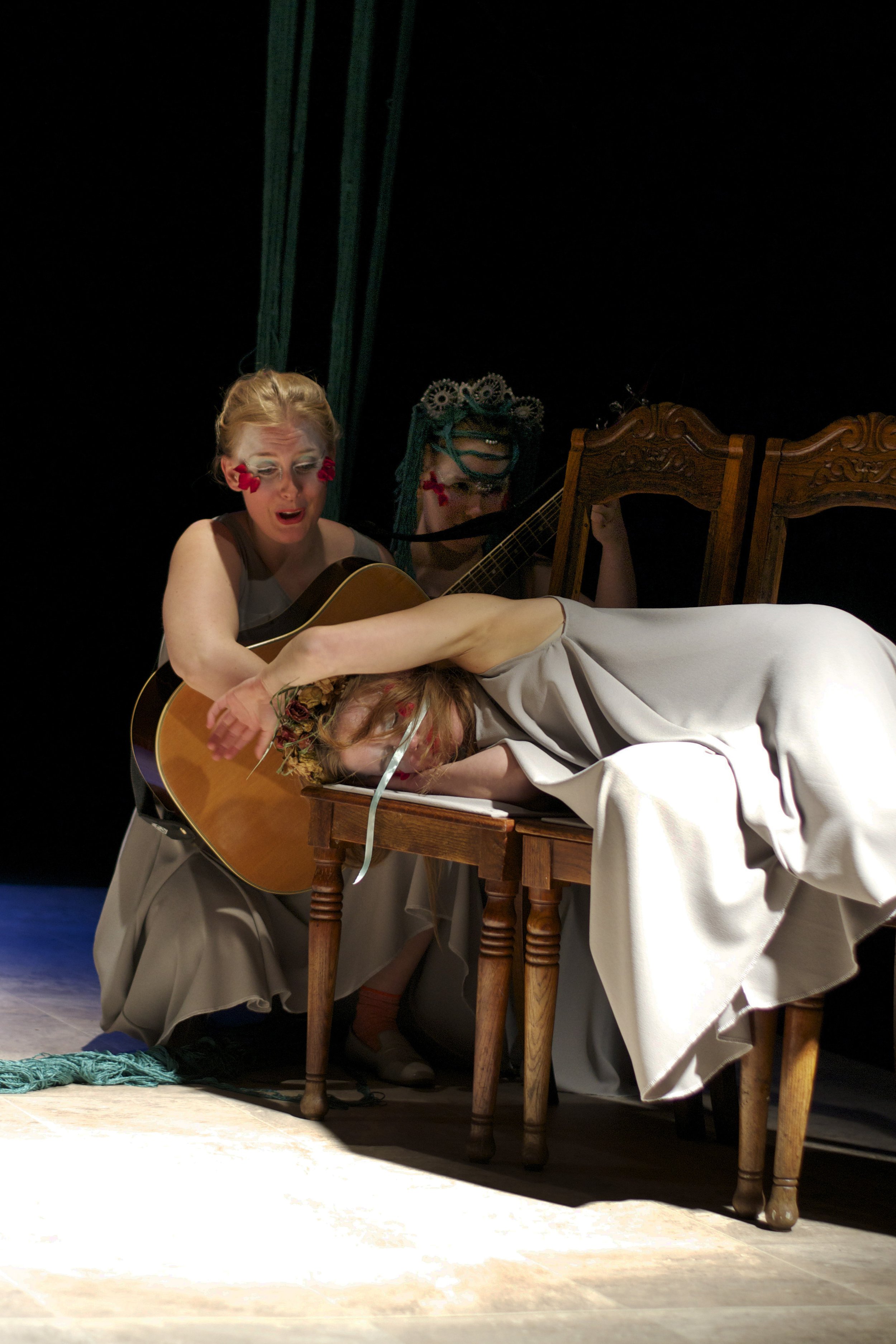 Three women in theatrical costumes and makeup are on stage; one plays a guitar, another leans over a table, and the third observes from behind.