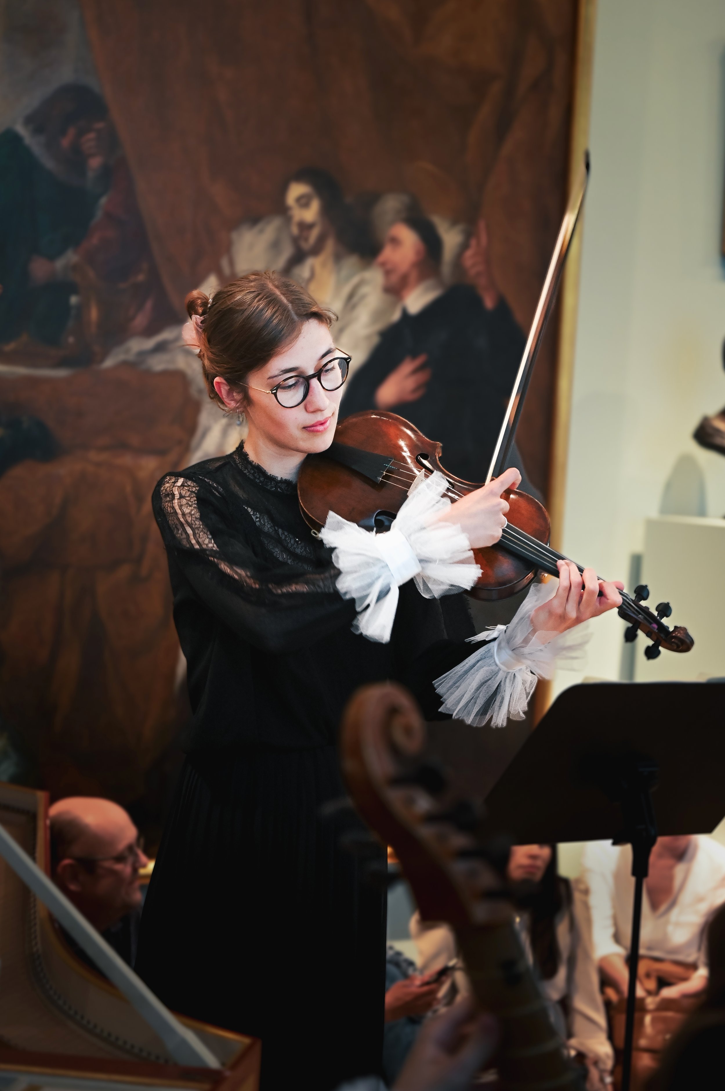 Woman in glasses playing a violin in front of a classical painting at an indoor event.