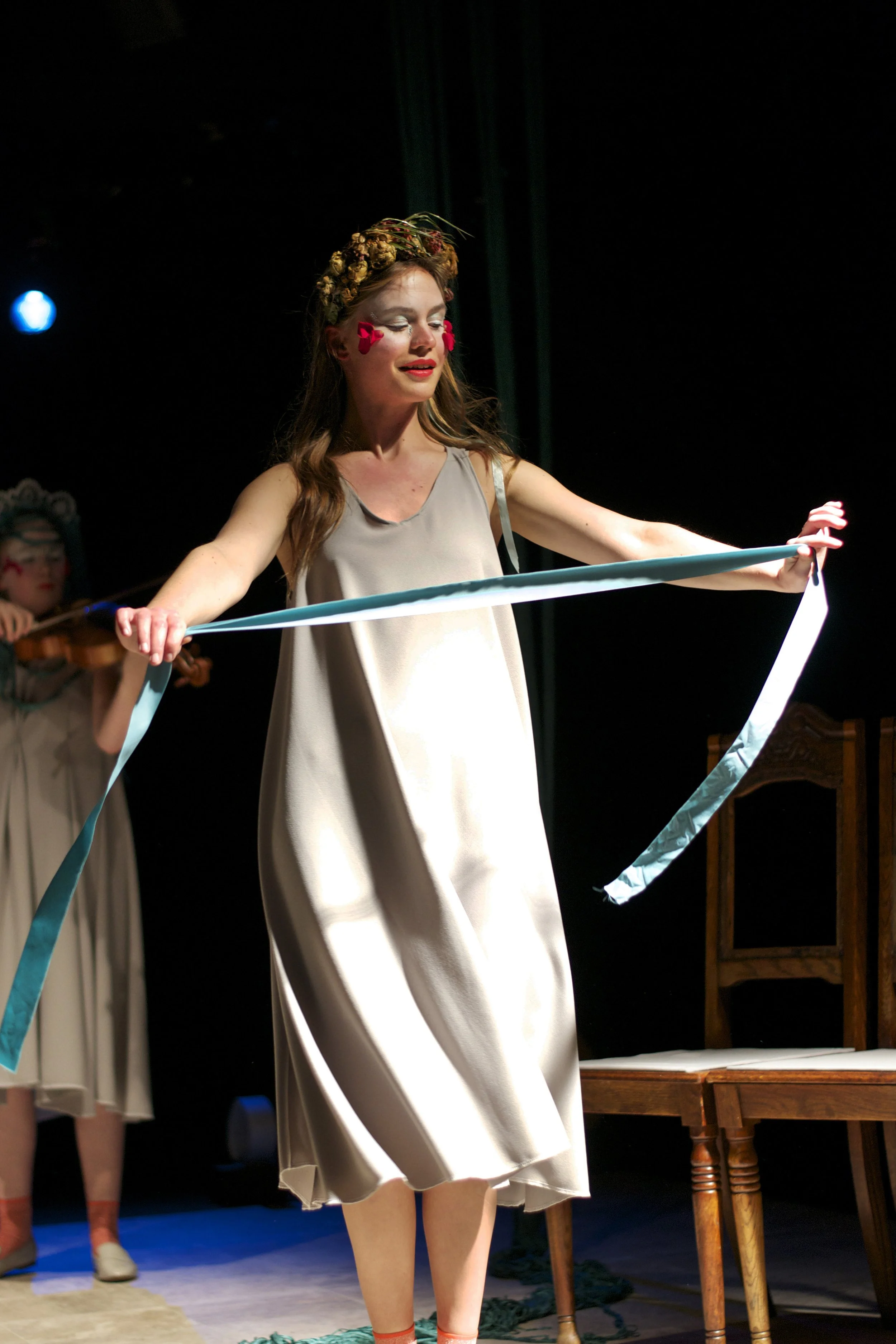 A performer on stage wearing a beige dress with face paint and a floral crown, holding a blue ribbon with outstretched arms. Another performer in similar attire plays a violin in the background.