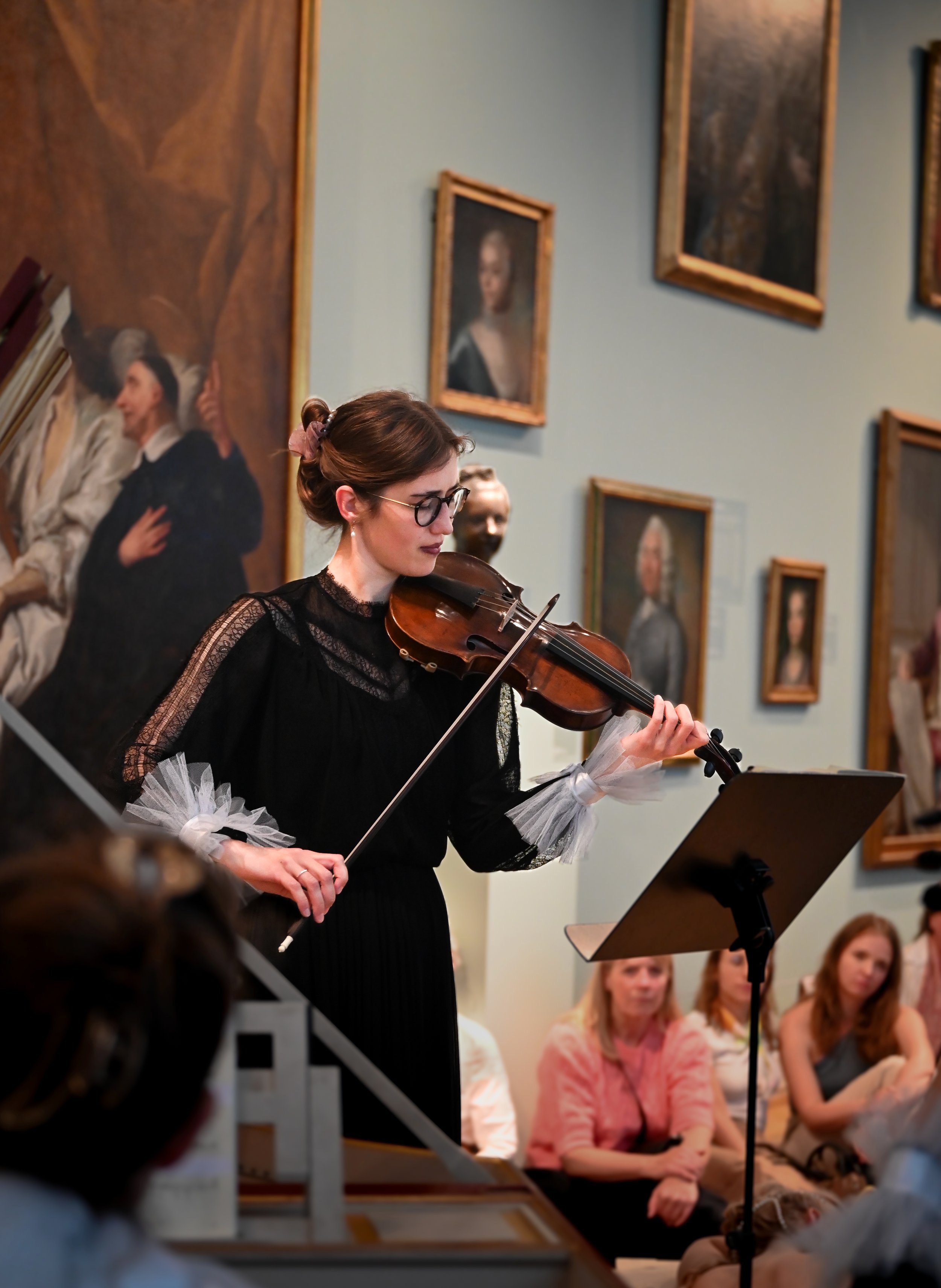 Woman playing violin in an art gallery surrounded by paintings, with an audience watching.