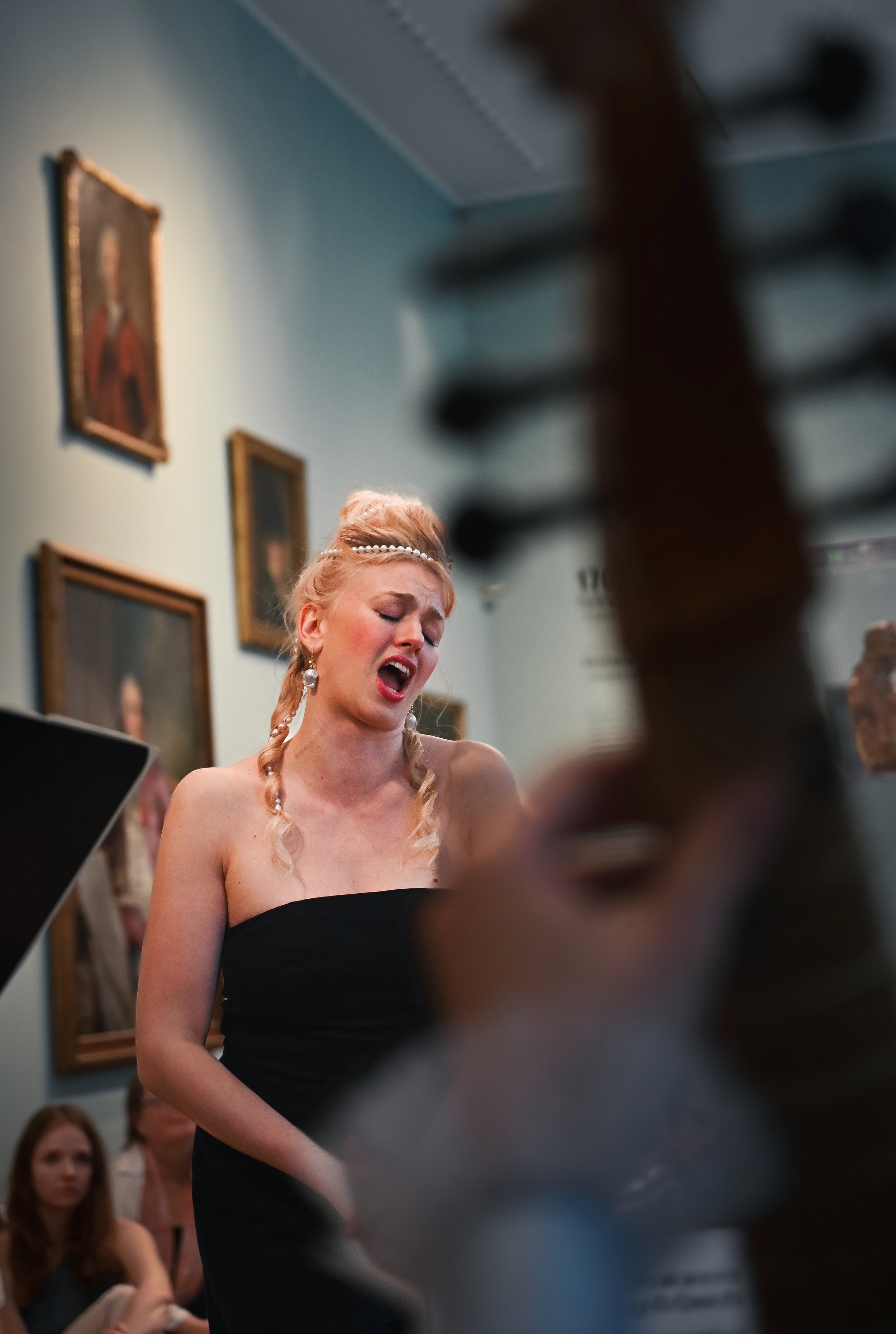 A woman singing passionately in a room with framed paintings on the walls, with a musical instrument's neck in the foreground.