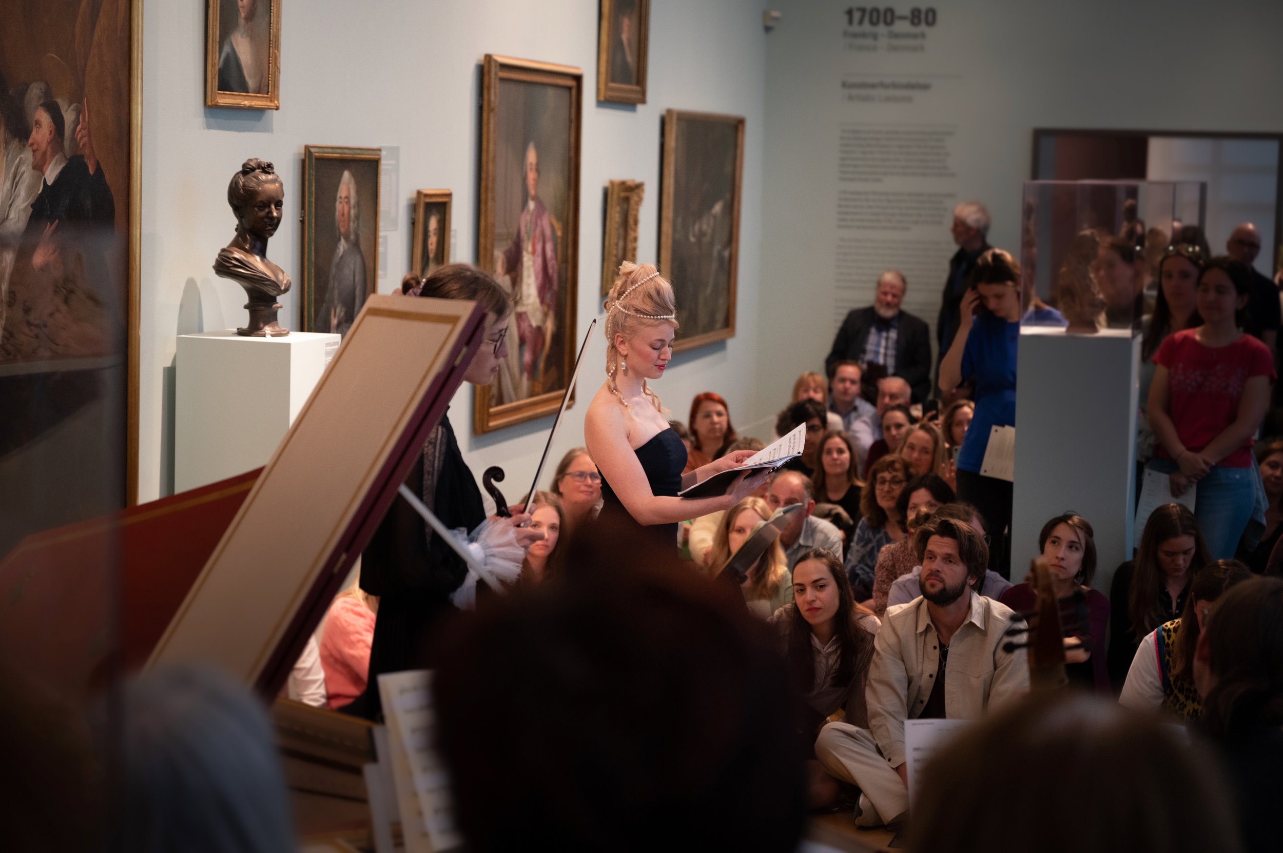 A woman in an elegant dress reads sheet music during a classical music performance at an art gallery. The audience is seated and standing, surrounded by framed paintings and sculptures. The atmosphere is attentive and cultured.