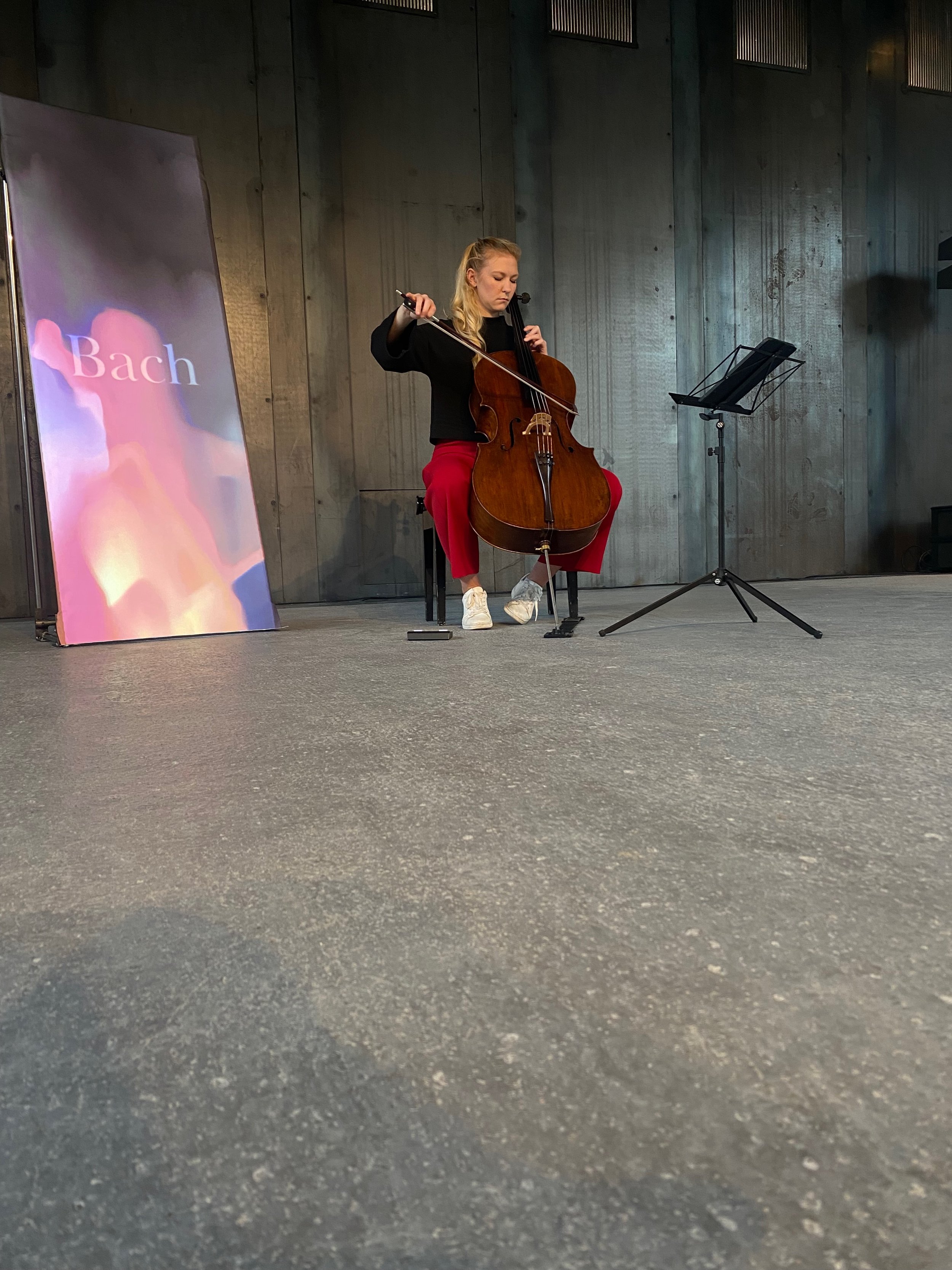A musician playing a cello next to a sign labeled 'Bach', in a performance setting with a music stand.