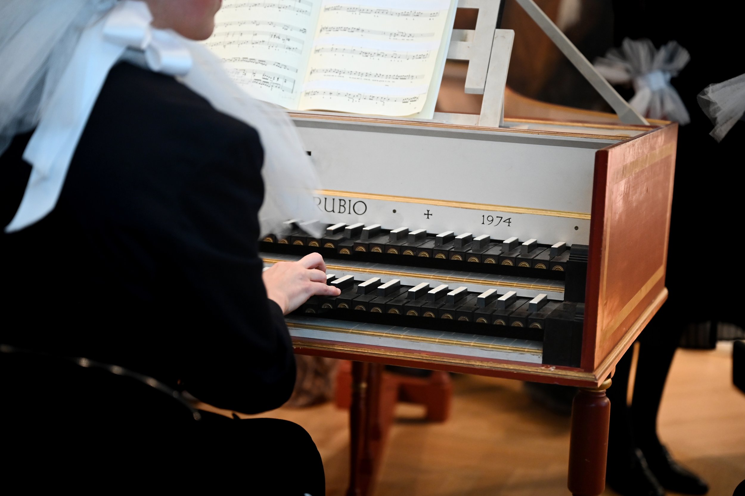 Person playing a harpsichord with sheet music in the background, wearing formal attire with a bow.