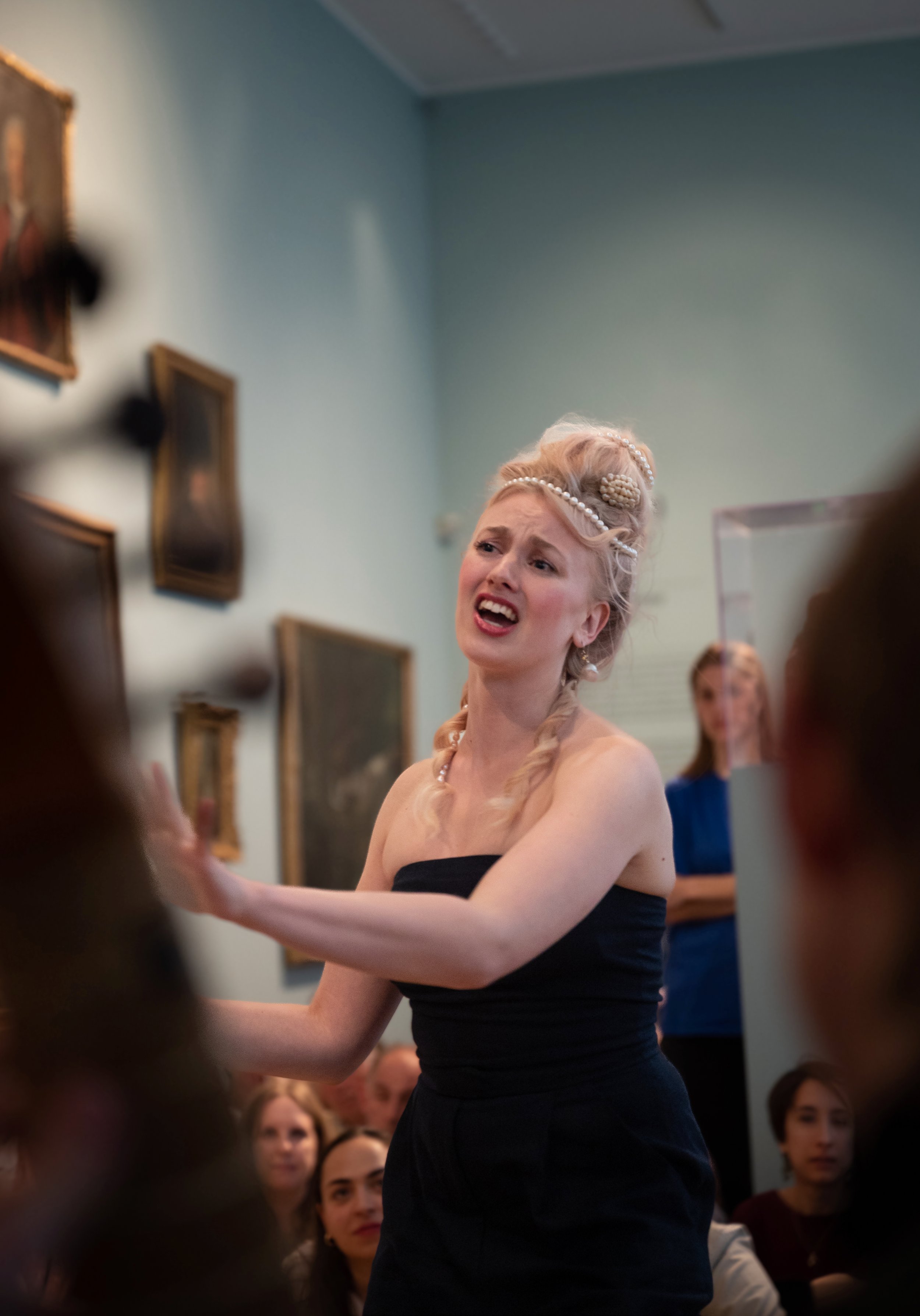 A woman wearing a black strapless dress is passionately performing in front of an audience in an art gallery. She has elaborate, styled hair adorned with pearls and appears to be singing or speaking dramatically. Several paintings are visible on the