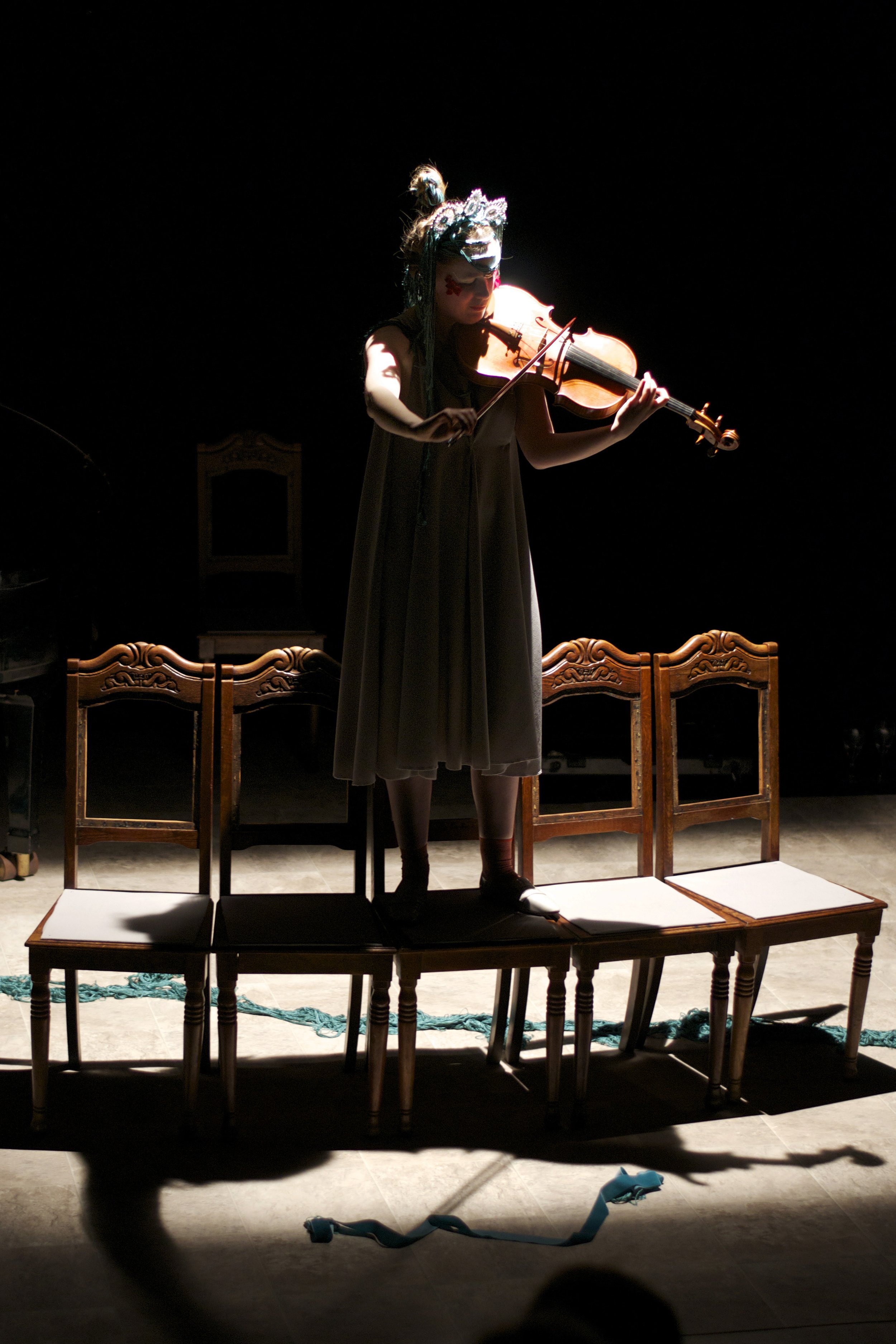 A person wearing a mask and a decorative headpiece plays the violin while standing on a row of chairs in a dimly lit room. There is a shadow cast on the floor and a rope or ribbon is visible in the background.