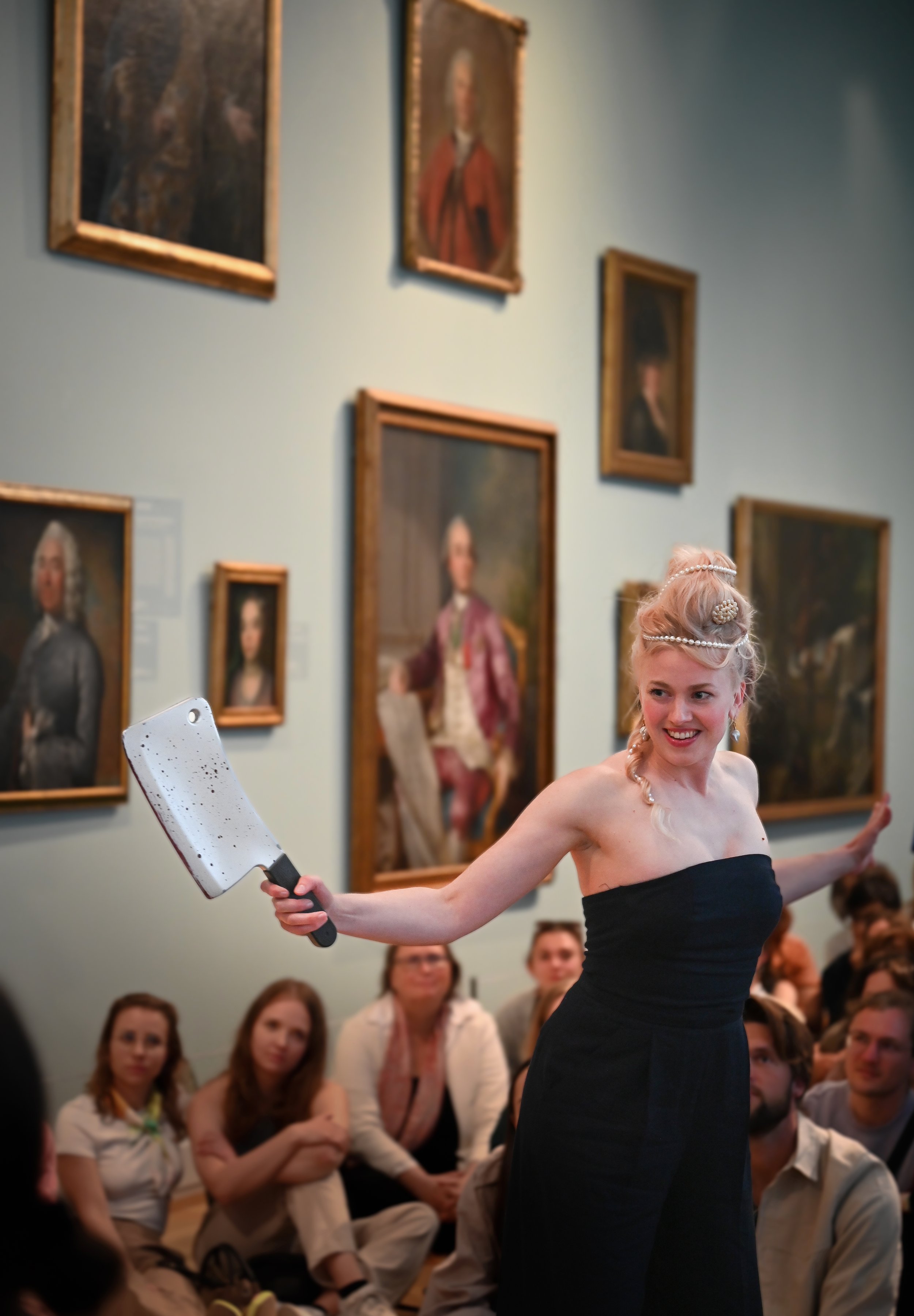 A woman in a black strapless dress posing with a meat cleaver in front of a seated audience, in an art gallery with classic portraits on the walls.