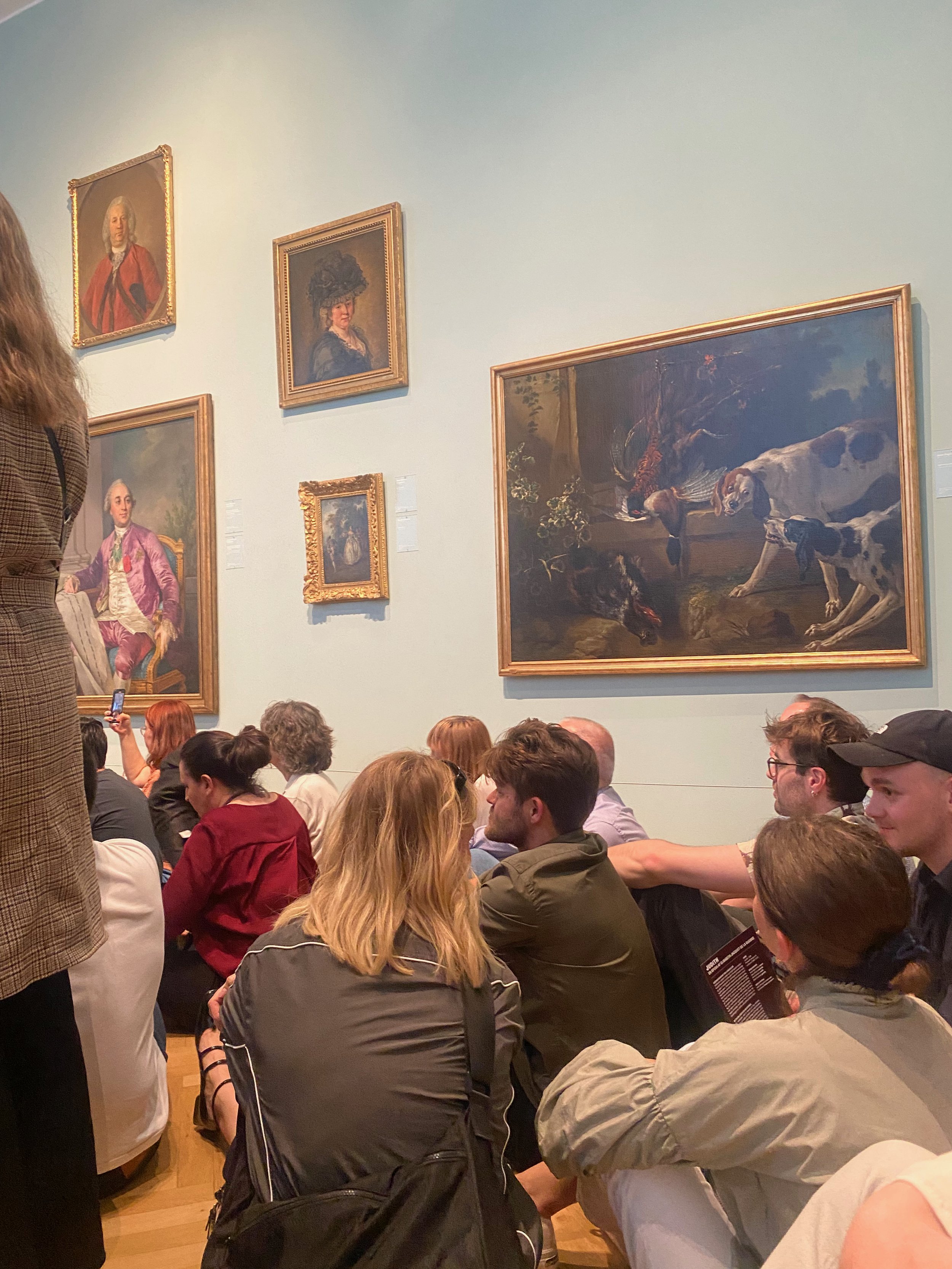 A group of people sitting on the floor of an art gallery, looking at several classic paintings on the wall. The gallery features portraits and a painting of hunting dogs. Some people are sitting cross-legged and appear to be listening to a presentati