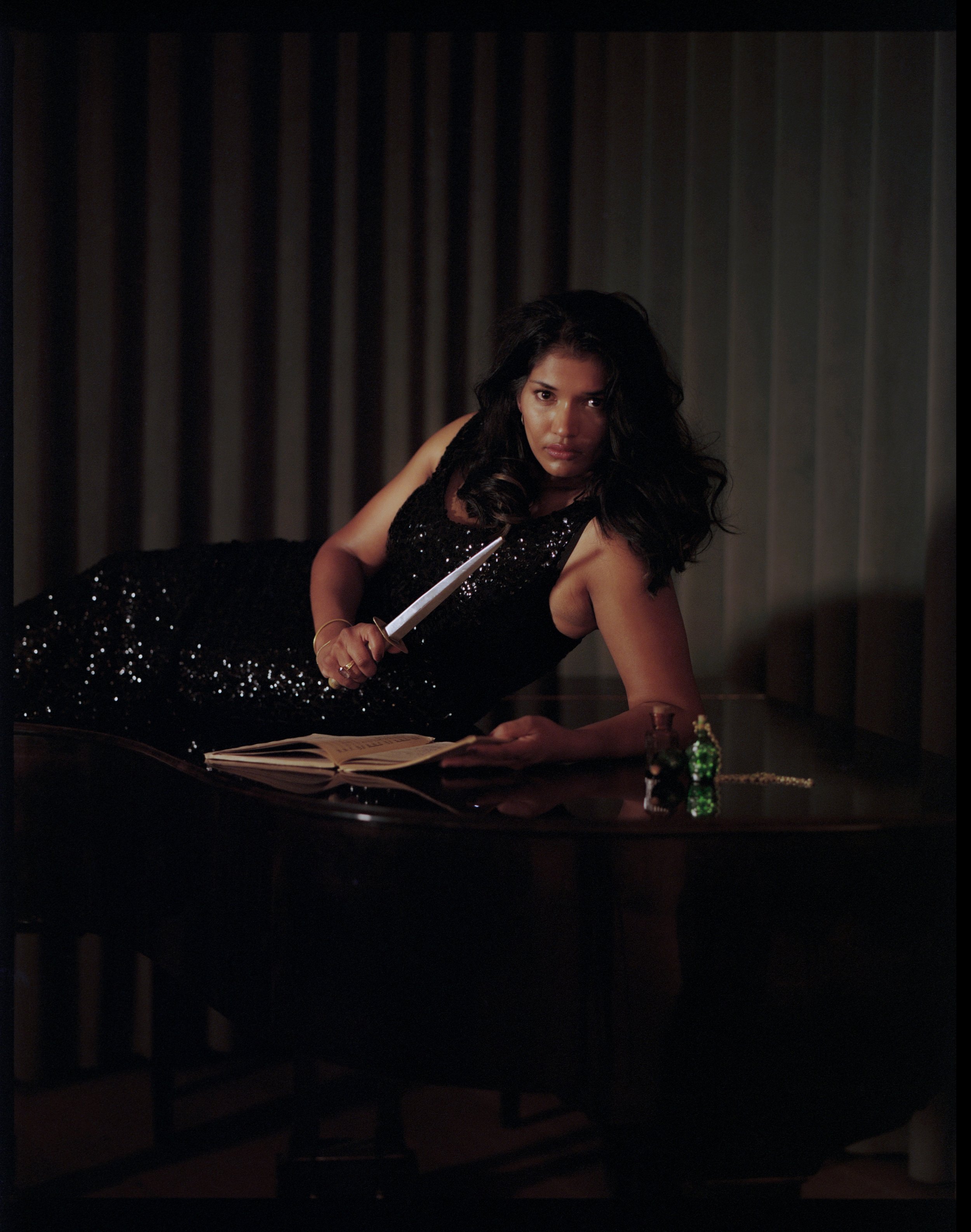 A woman in a black sequined dress is reclining on a piano, holding a pen or writing instrument over an open book. There are small decorative items like a green bottle and a string of pearls on the piano.