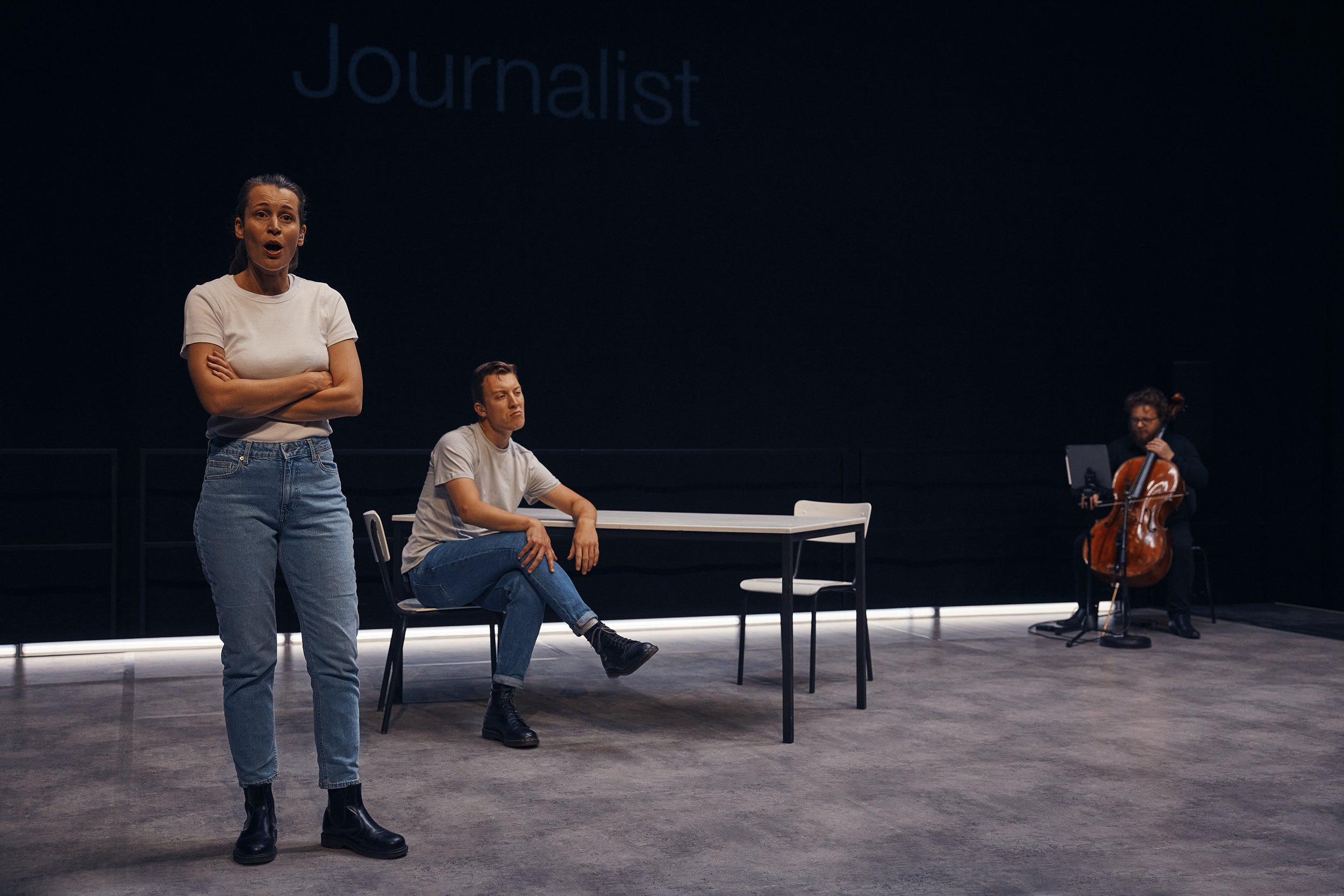 Three people in a black box theater setting: a person standing and speaking, a person sitting at a table, and a cellist playing music to the right.