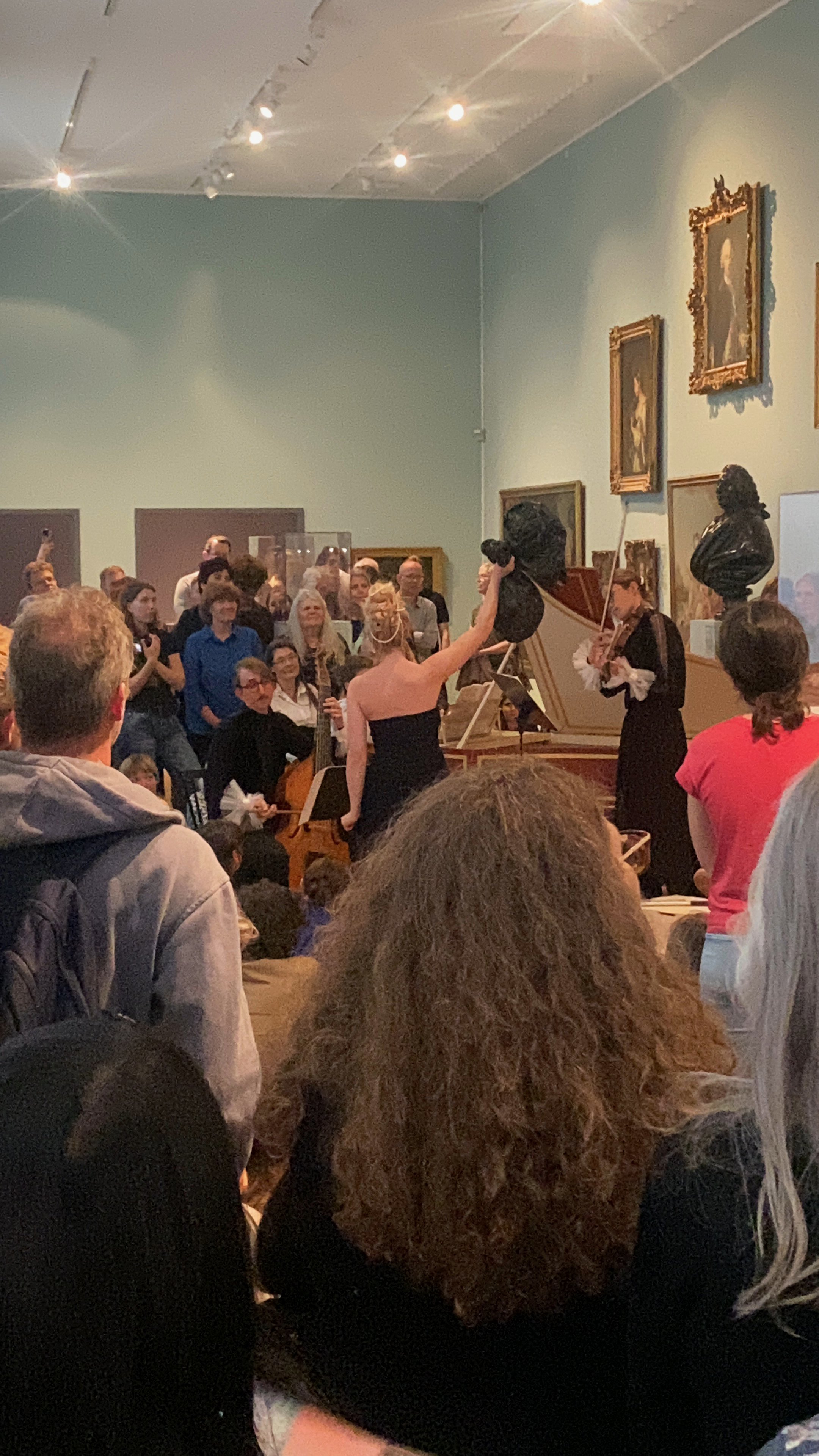 A crowded interior room with people gathered around musicians in front of art pieces and sculptures.