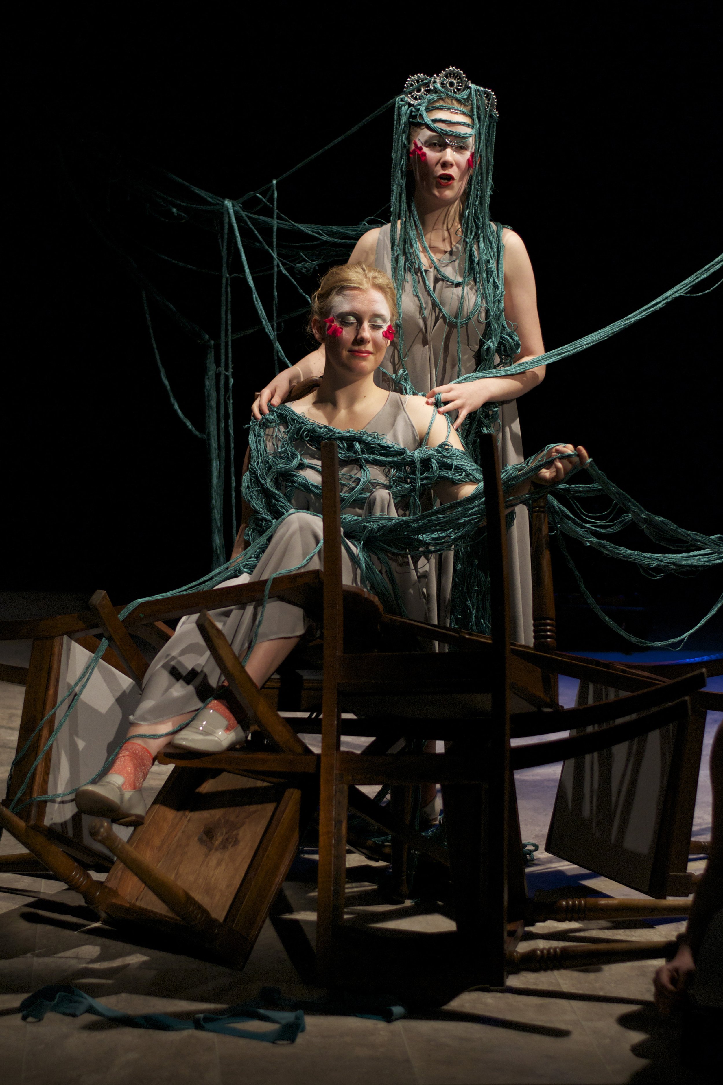 Two women on stage in theatrical costumes, surrounded by tangled blue yarn and wooden chairs. One is seated with eyes closed and the other stands behind her, both wearing dramatic makeup with red accents.