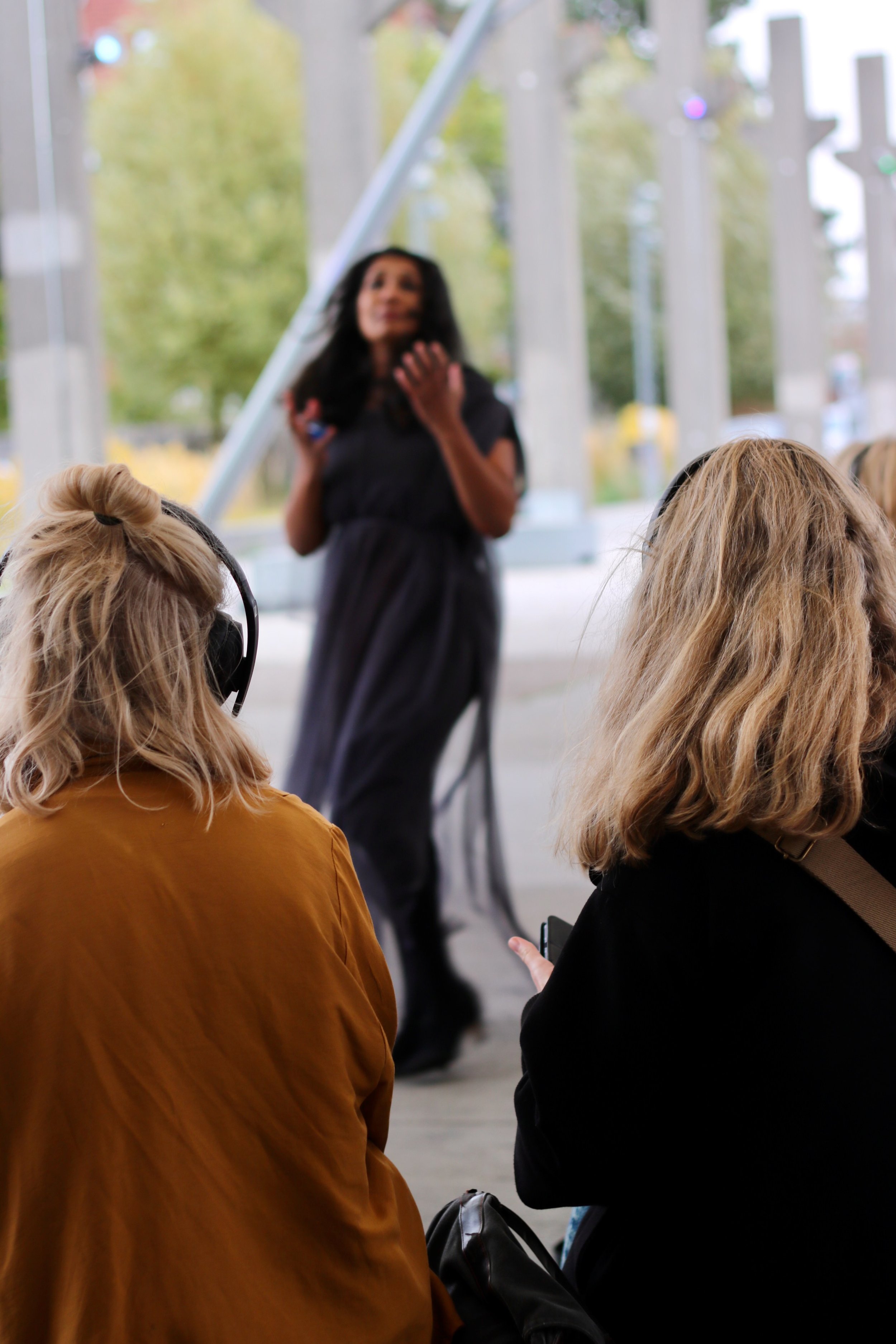 Two women with headphones listen to a speaker in an outdoor setting with trees and columns in the background.
