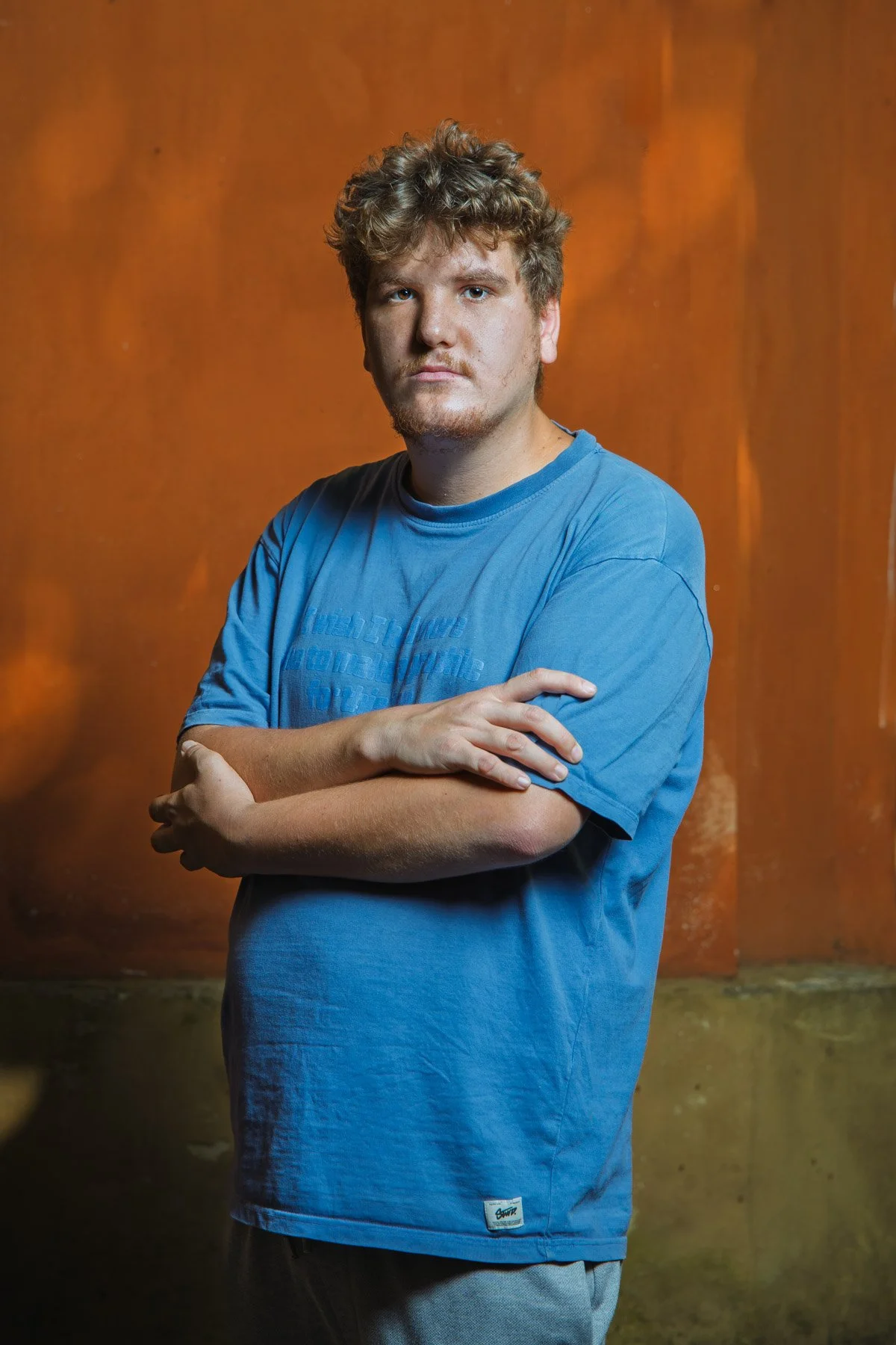 Ragazzo con felpa blu che posa con le braccia incrociate di fronte a uno sfondo arancione.