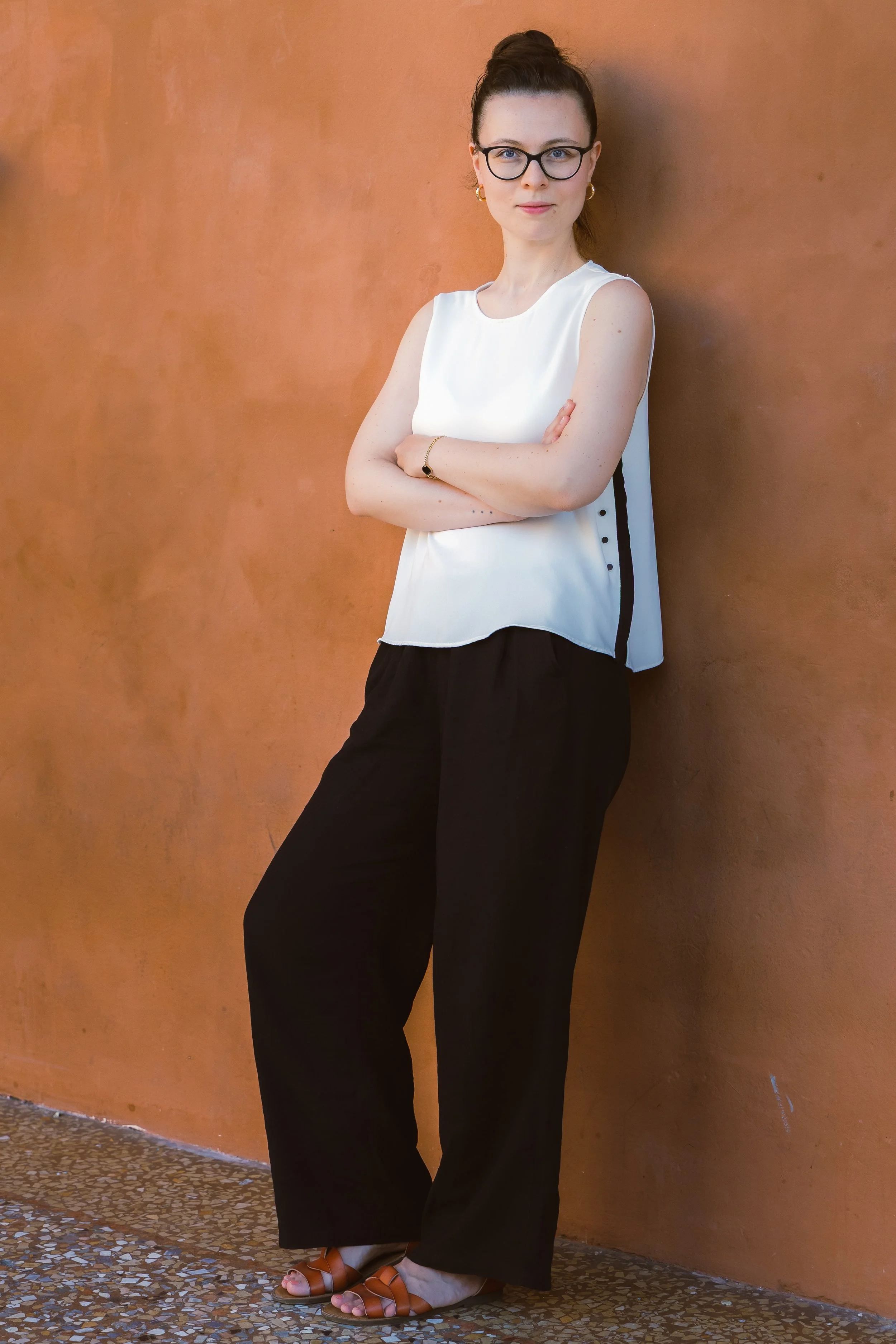 Una donna con occhiali, capelli raccolti, indossa una maglietta senza maniche bianca e pantaloni larghi neri, si appoggia a una parete arancione.