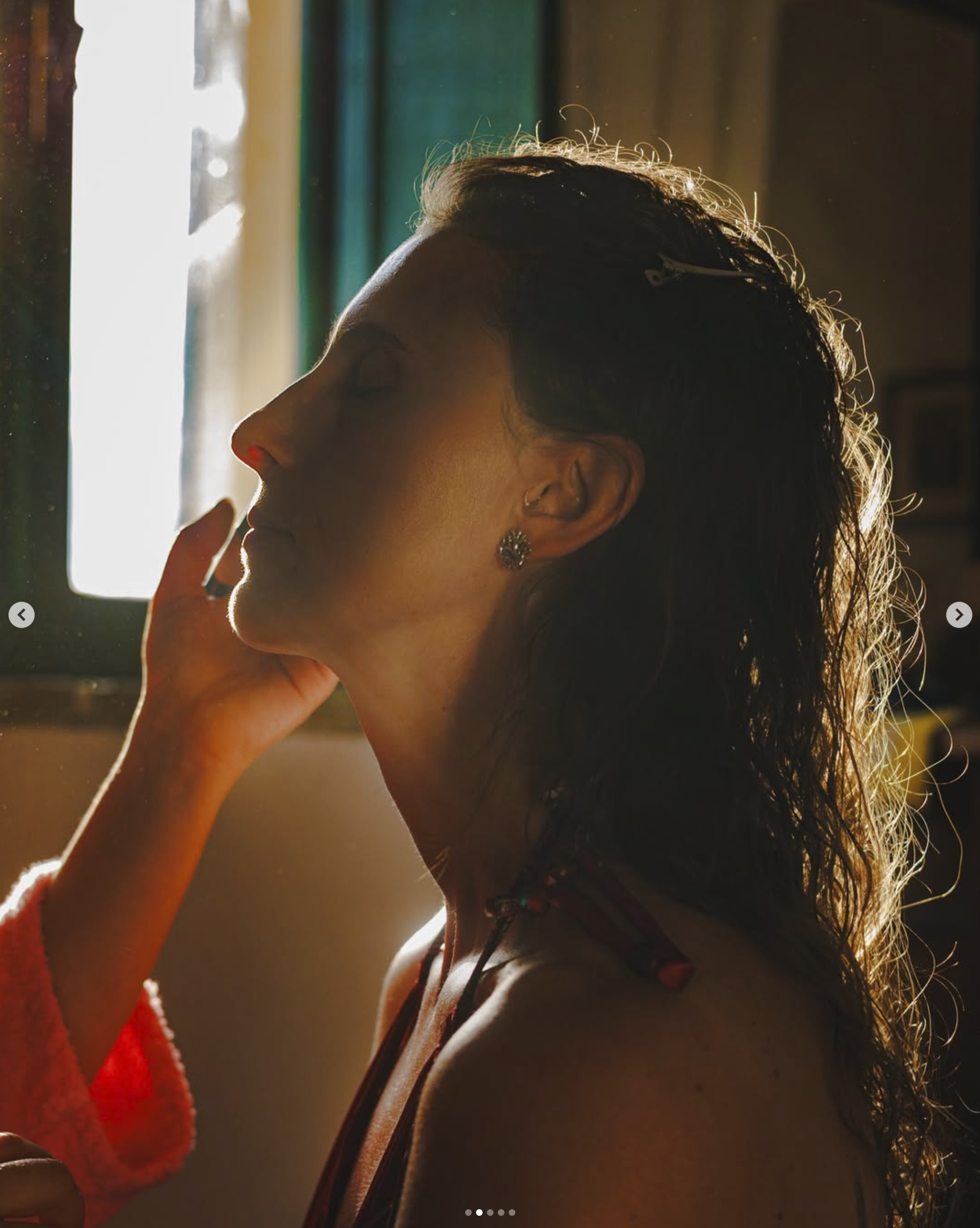Ragazza con capelli ricci scuri che si riposa con gli occhi chiusi, illuminata dal sole vicino a una finestra, con orecchino pendente e collana.