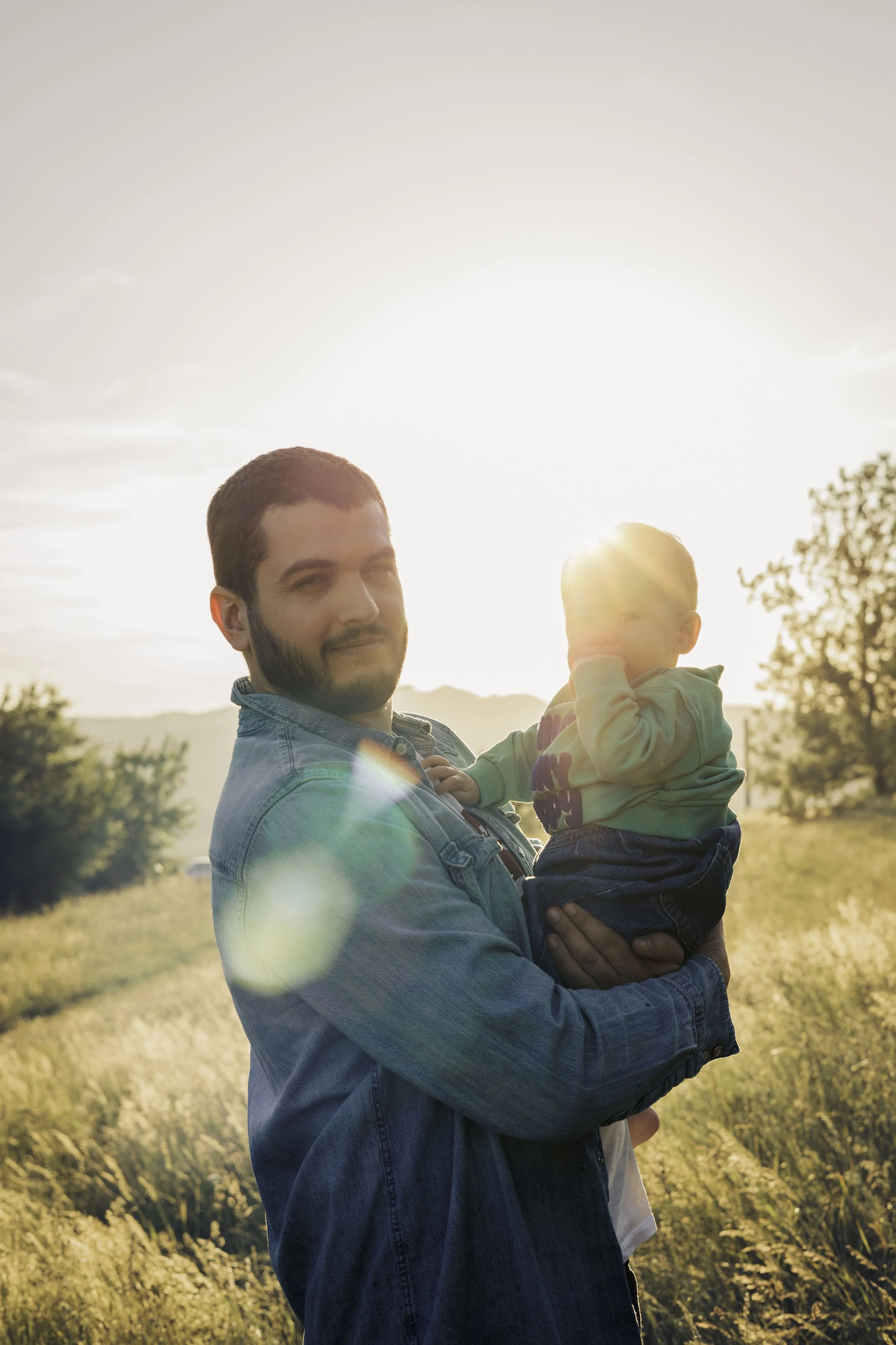 Un uomo tiene un bambino in braccio in un campo al tramonto, con il sole che splende dietro di loro.