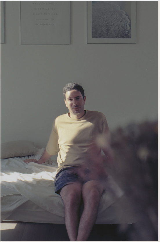 Uomo seduto sul letto in una stanza ben illuminata dalla luce naturale, con due quadri sulla parete alle sue spalle.