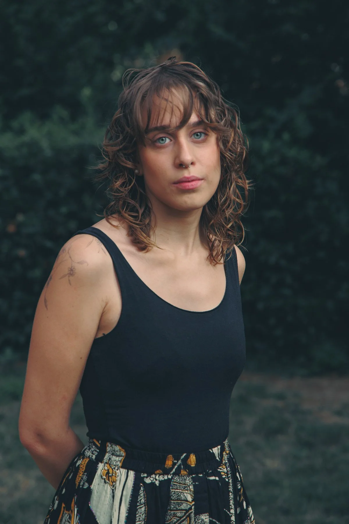 Ragazza con capelli ricci e occhi chiari, indossa una maglietta nera senza maniche e una gonna colorata a fantasia, si trova all'aperto in un'area verde e naturale.
