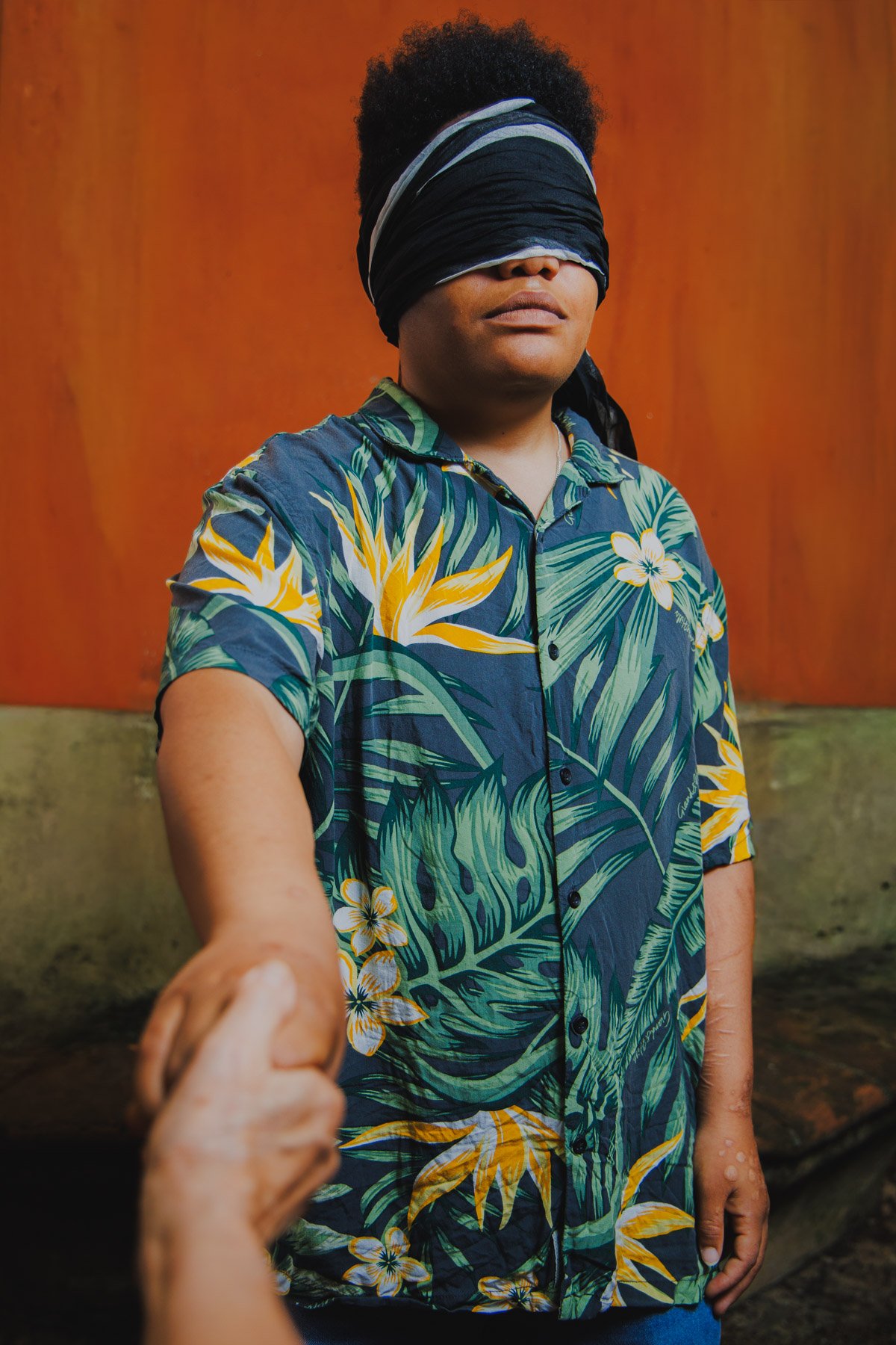 Donna con occhiali appoggiati sugli occhi, indossa una camicia con stampa tropicale di foglie e fiori di colore verde, giallo e bianco, mentre tiene la mano di un'altra persona.
