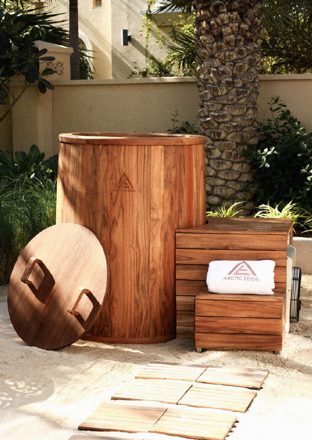 Outdoor scene with wooden sauna, wooden stool with Arctic Edge logo, rolled towel, round wooden table with handles, sand, and stepping stones in a tropical garden.