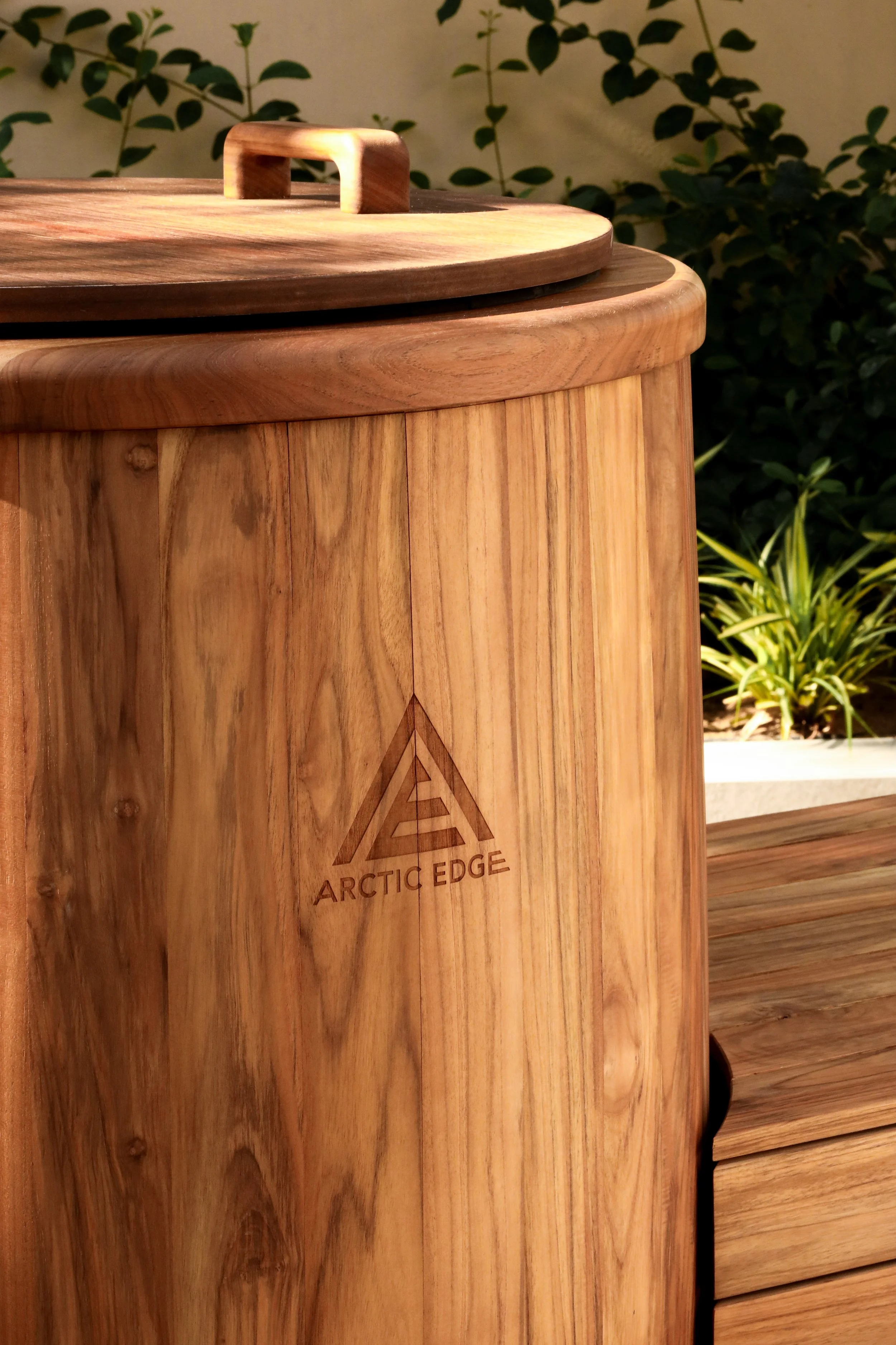 Close-up of a wooden container with a lid, engraved with the Arctic Edge logo, outdoors with plants in the background.