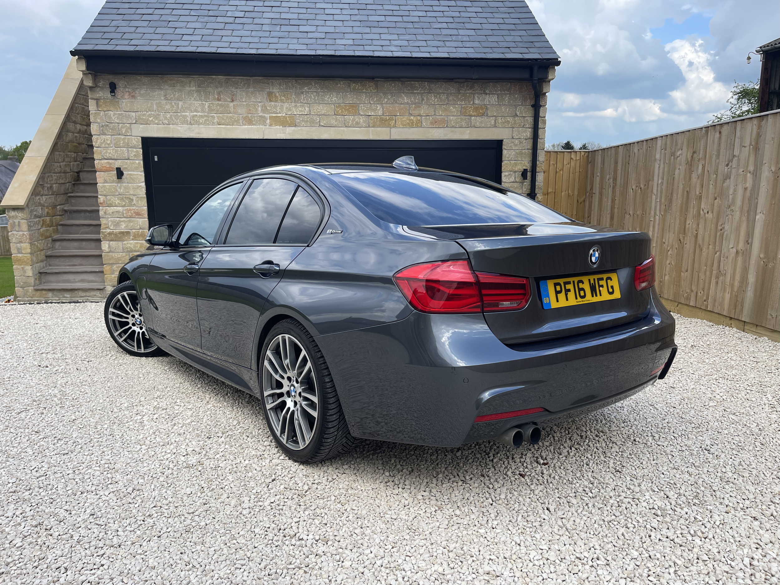 Gray BMW 3 Series sedan parked on gravel driveway, near a stone garage with black door.